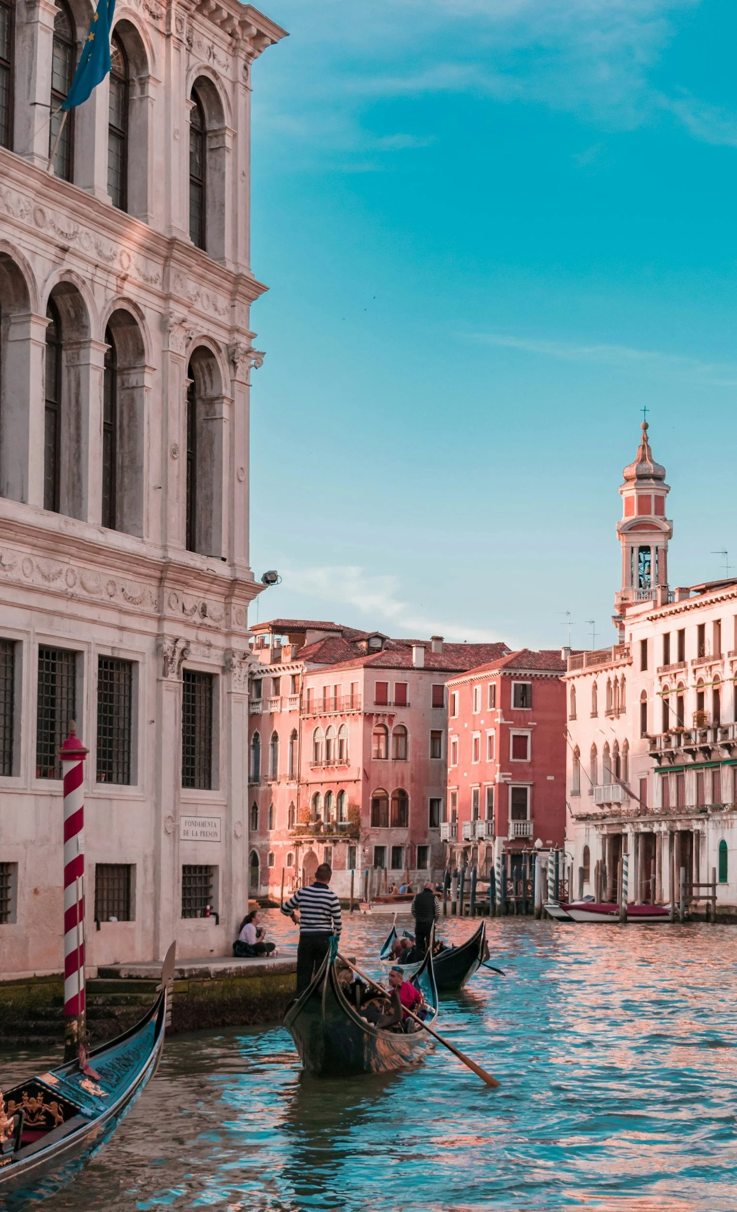 Gondolas floating on a canal in Venice, Italy, with historic buildings on either side and a blue sky above.