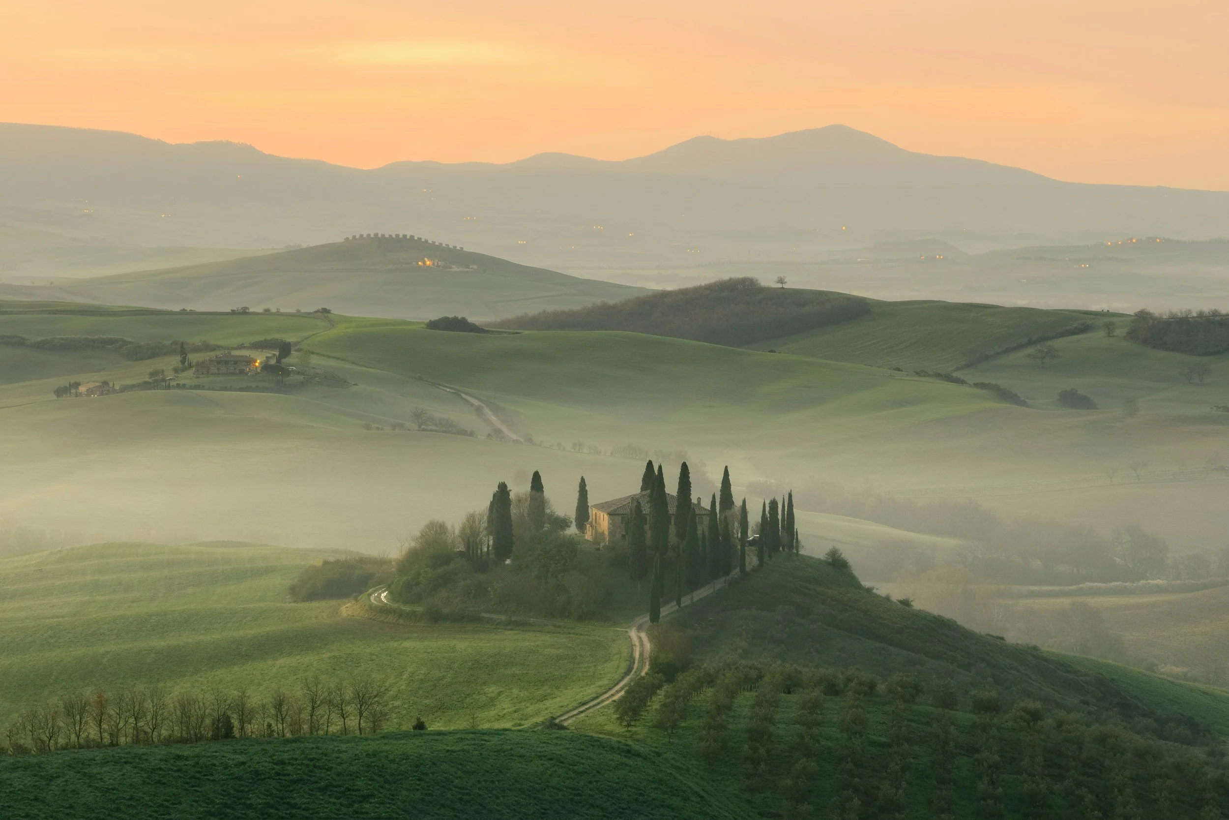 Landscape view of rolling green hills with a farmhouse surrounded by cypress trees, mist, and a pink-orange sky at sunrise or sunset.