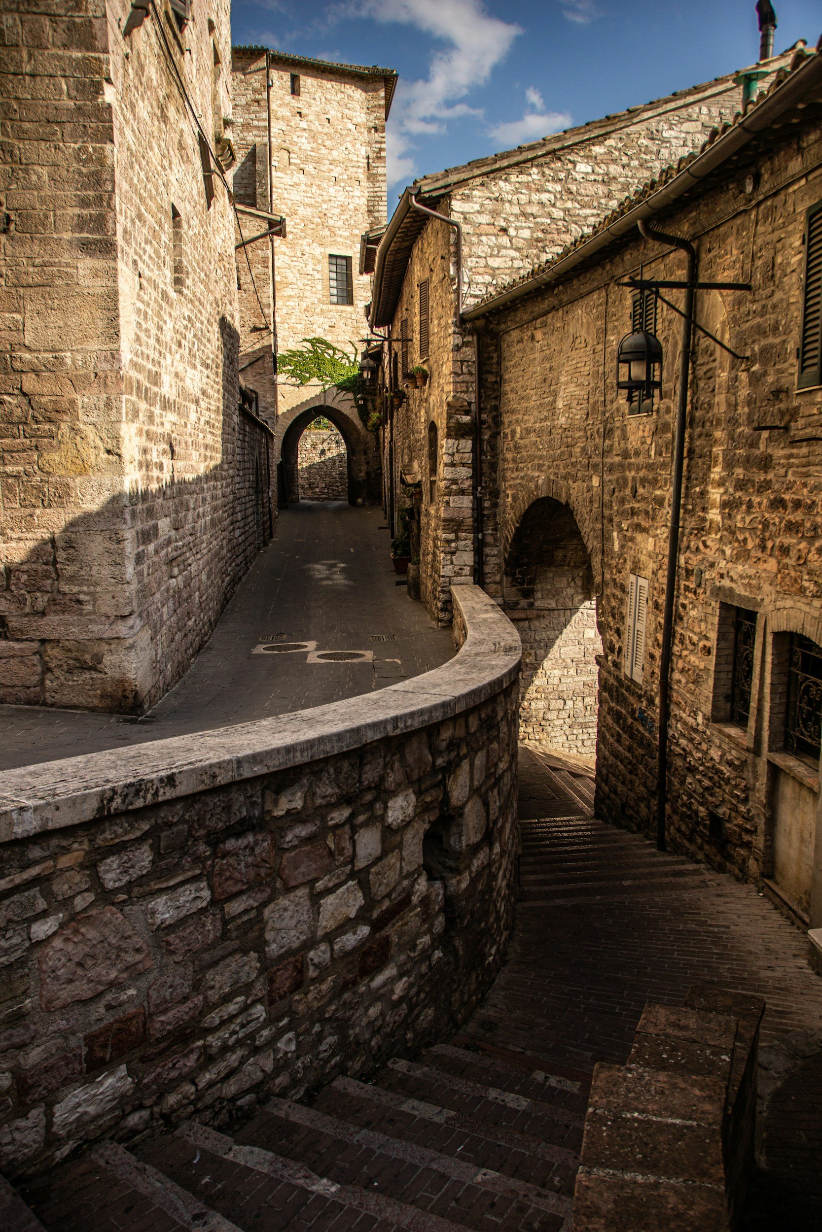 Narrow cobblestone street with stone buildings and archways in an old European town, bathed in warm sunlight with a blue sky and clouds overhead.