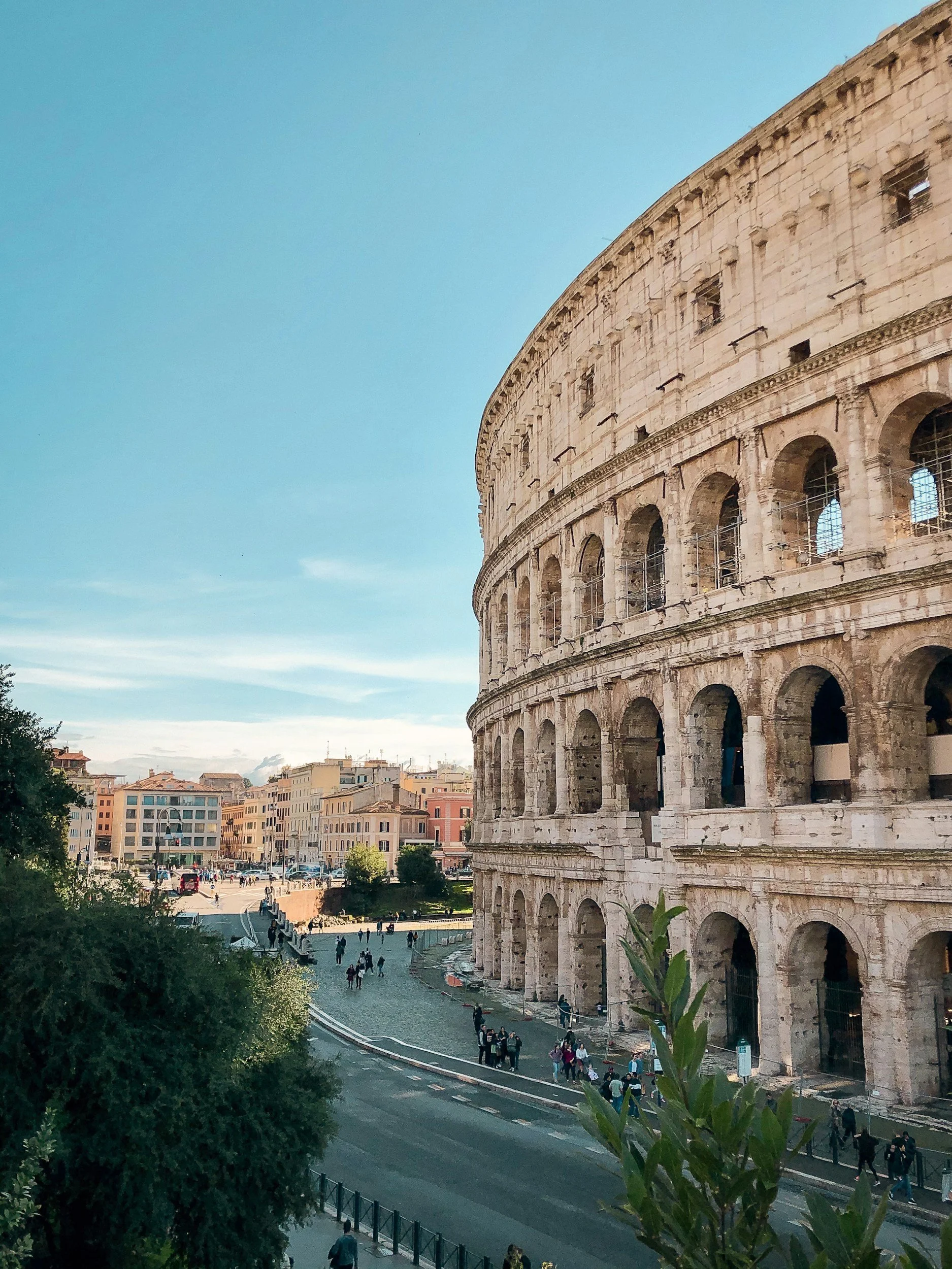 The image shows the Colosseum in Rome, Italy, with buildings and people nearby on a clear day.