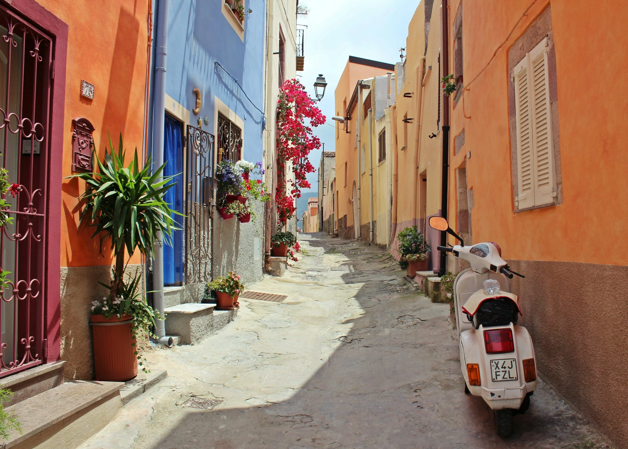 Narrow street lined with colorful houses with potted plants, a white scooter parked on the right, and flowers blooming along the street.