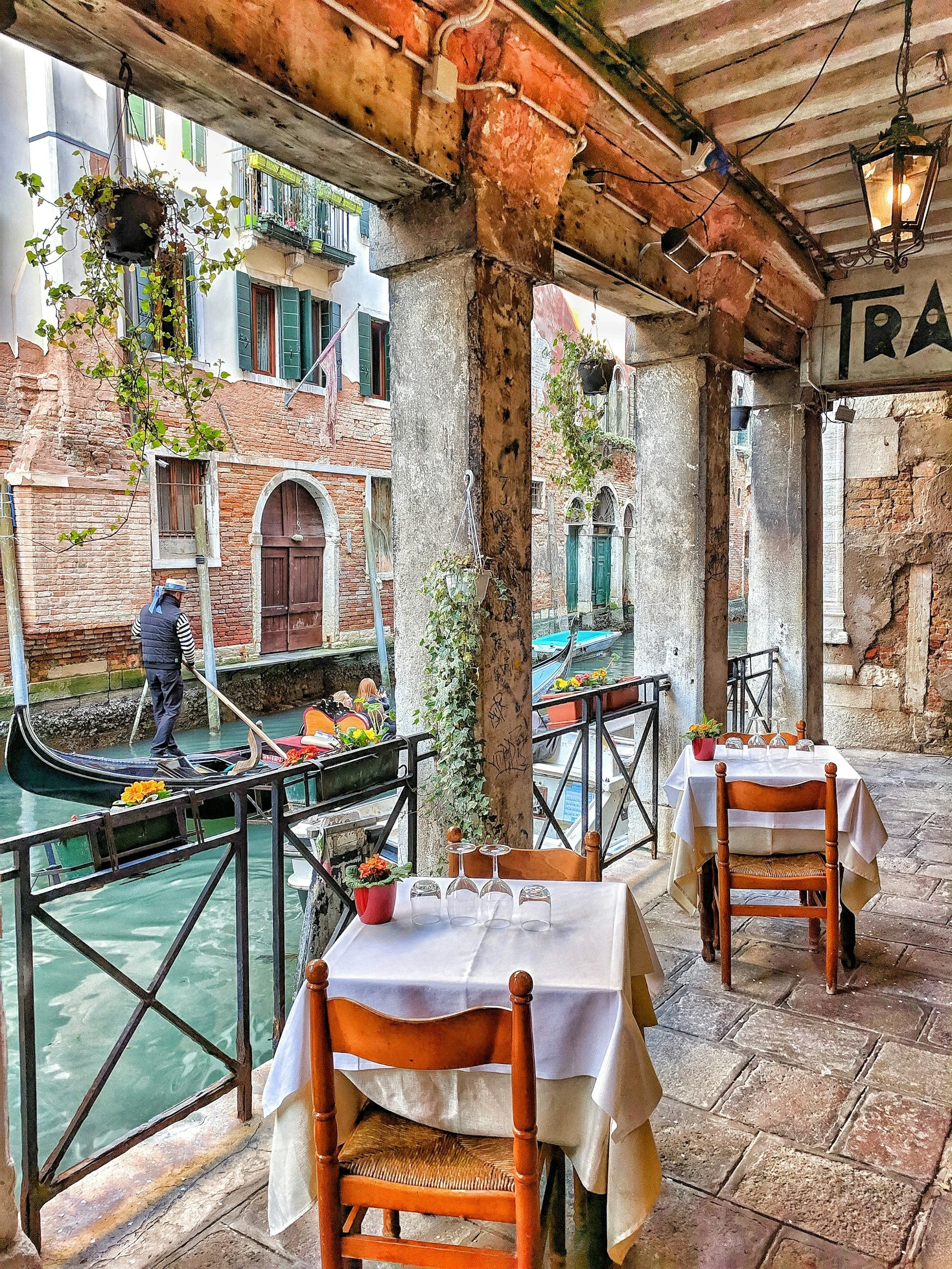 Outdoor cafe with white-clothed tables and chairs overlooking a canal with gondola boats passing by, in Venice, Italy.