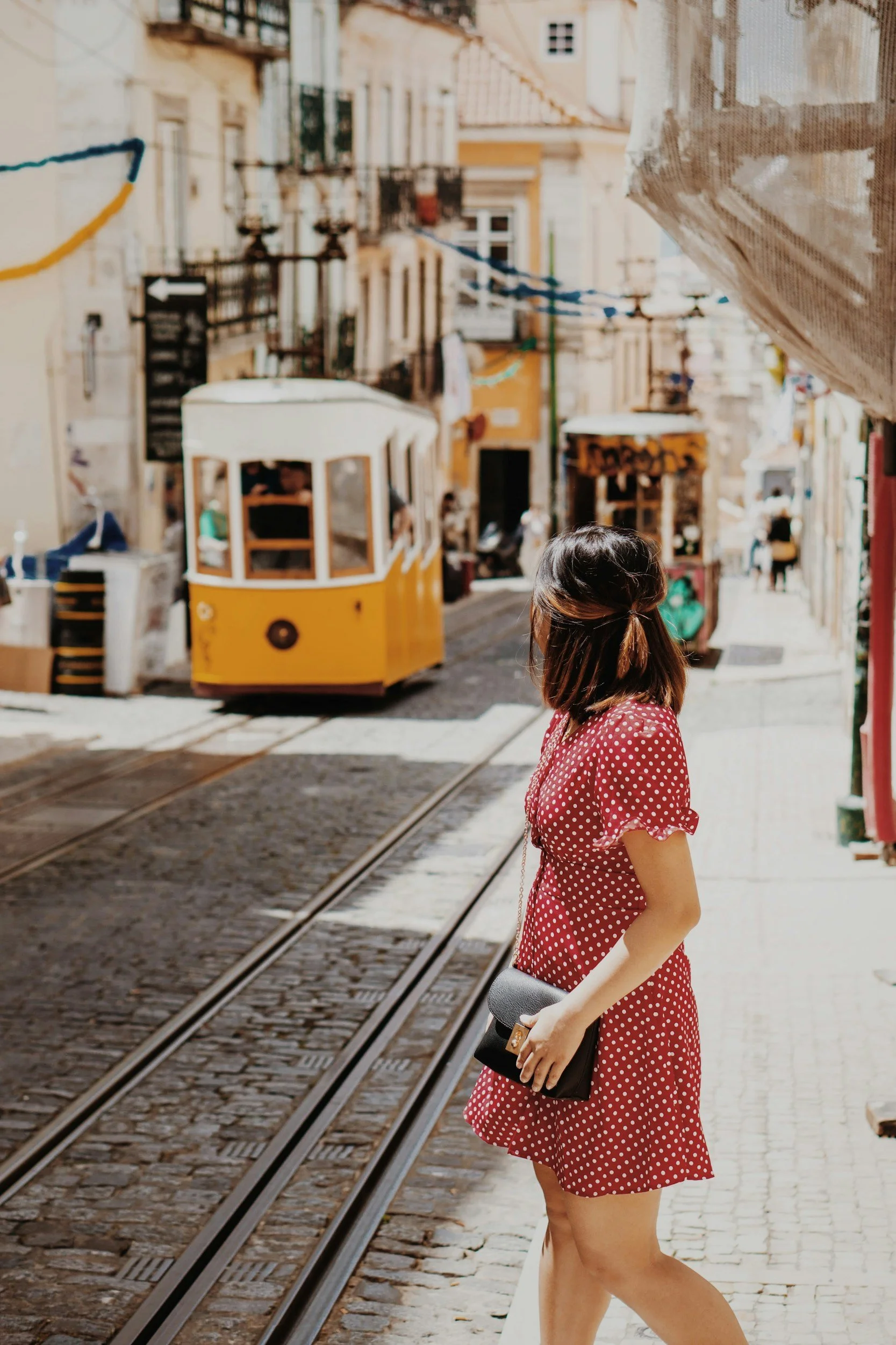 A woman with shoulder-length dark hair in a red polka-dot dress holding a black clutch purse, standing on a cobblestone street near tram tracks, looking toward a yellow tram in the background, with old buildings and string lights overhead.