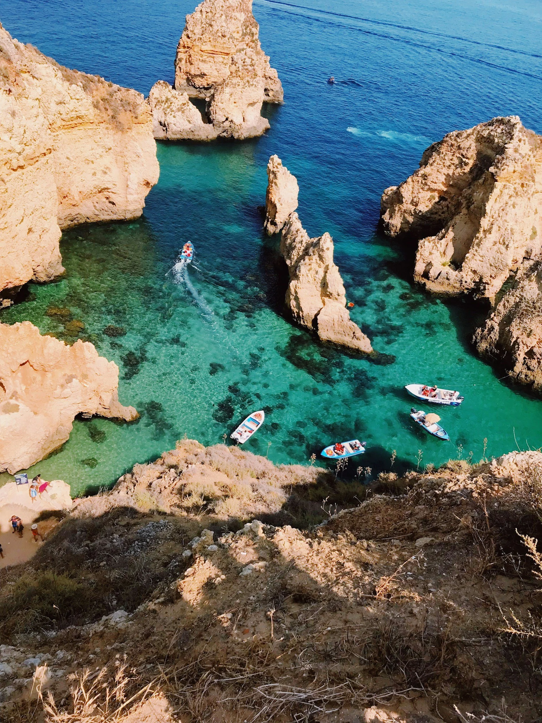 Aerial view of a turquoise cove with rocky cliffs, several boats floating, and swimmers near the shore.