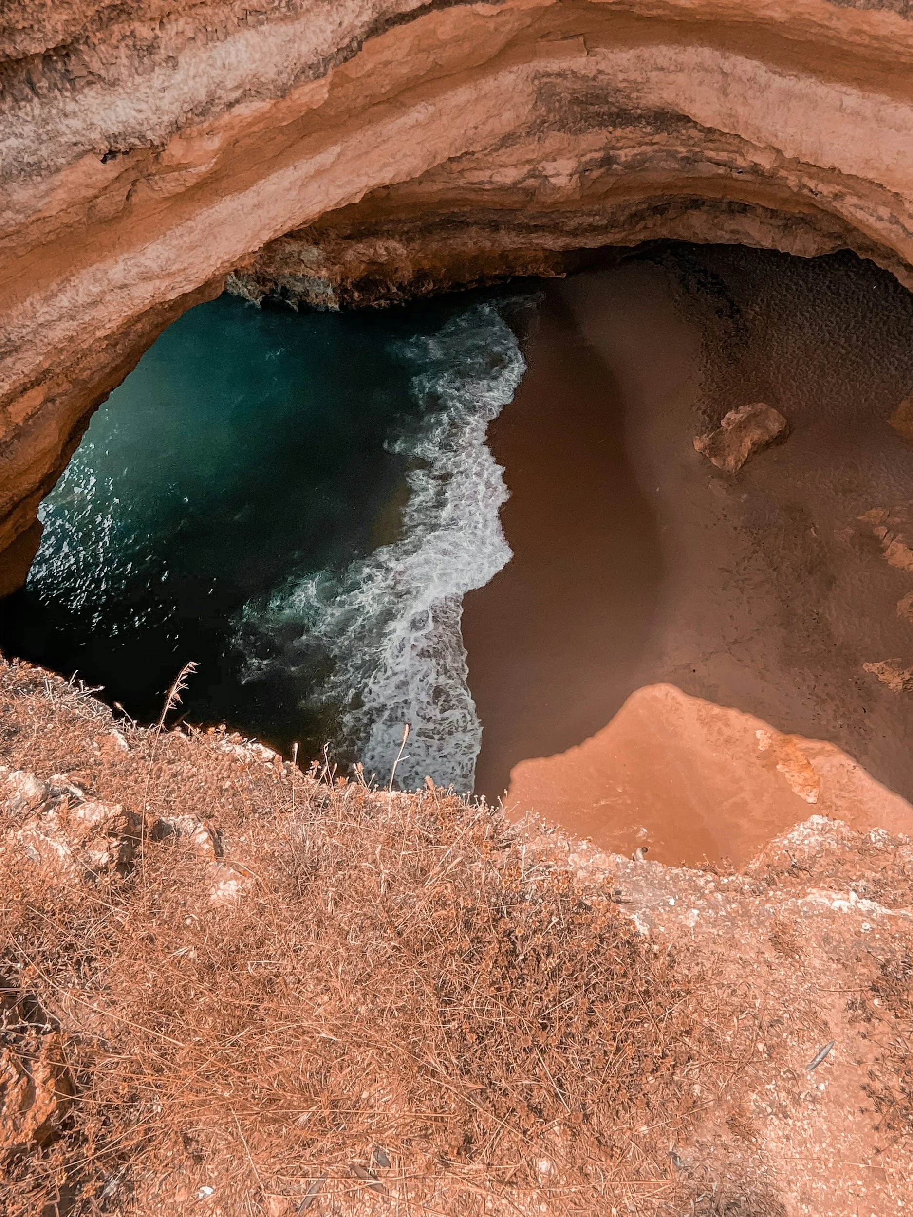 View of a natural sea cave with a sandy beach and ocean waves inside, surrounded by reddish-brown rocky cliffs.