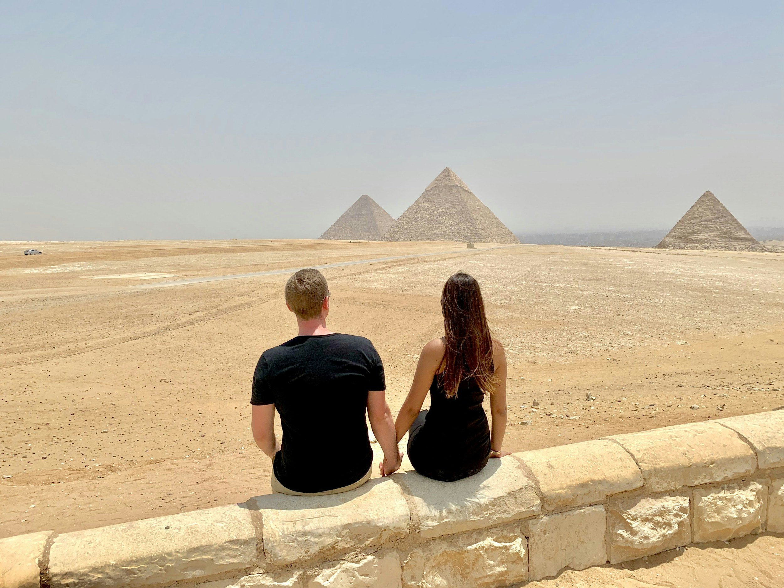A couple sitting on a stone ledge in a desert view of the pyramids of Egypt in the distance.