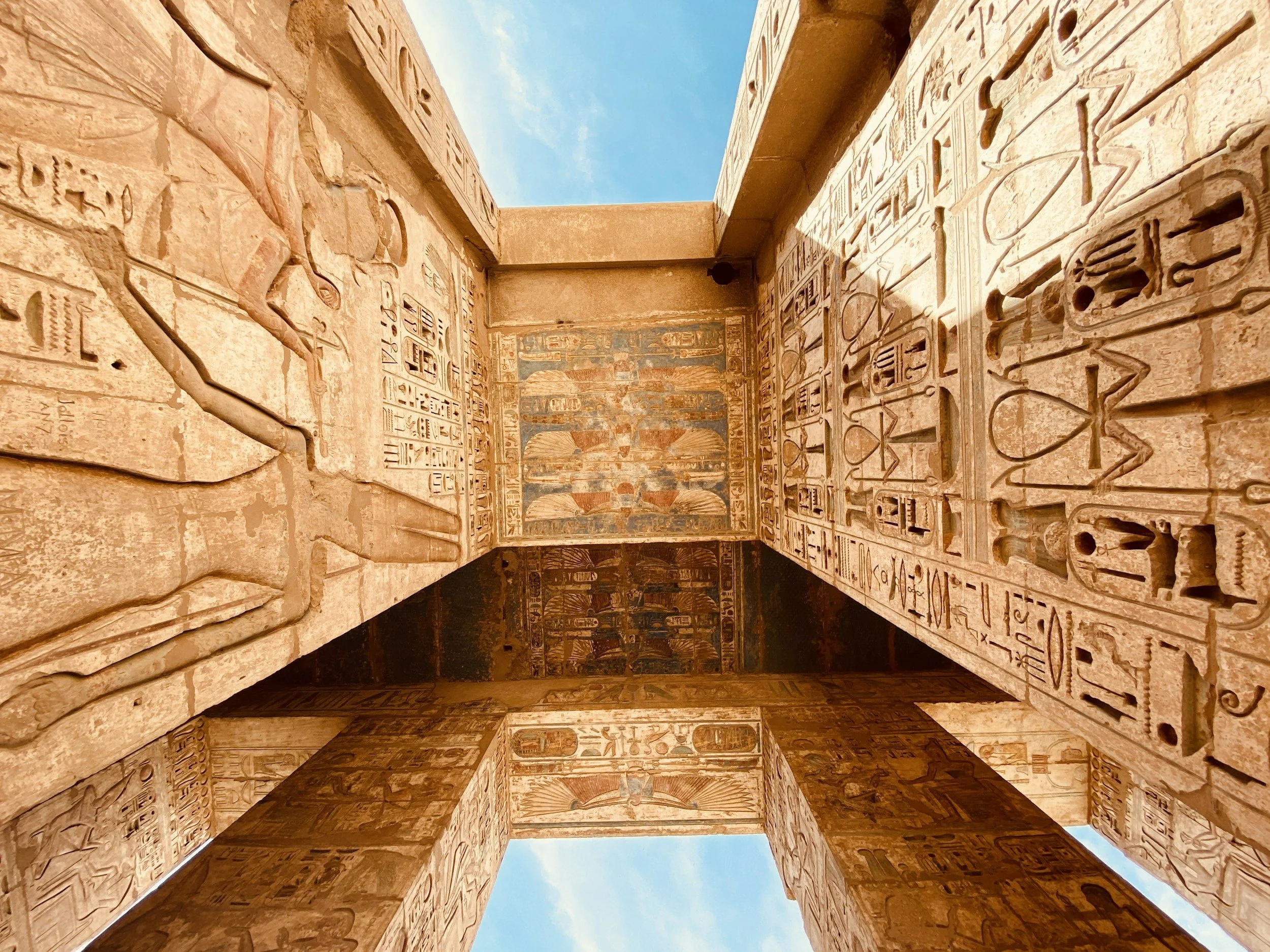 View of ancient Egyptian hieroglyphs and carvings on stone walls of a temple, with a clear blue sky visible above.