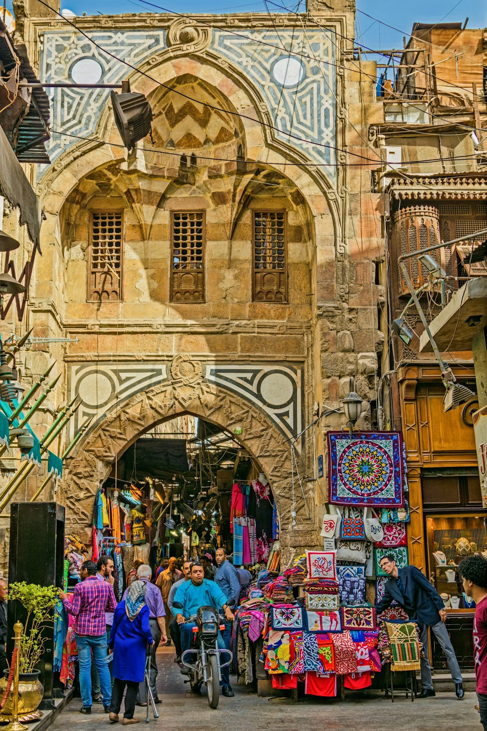 A busy marketplace street scene with a historic stone archway and building facade in the background, and various vendors selling colorful textiles, clothing, and souvenirs in the foreground.