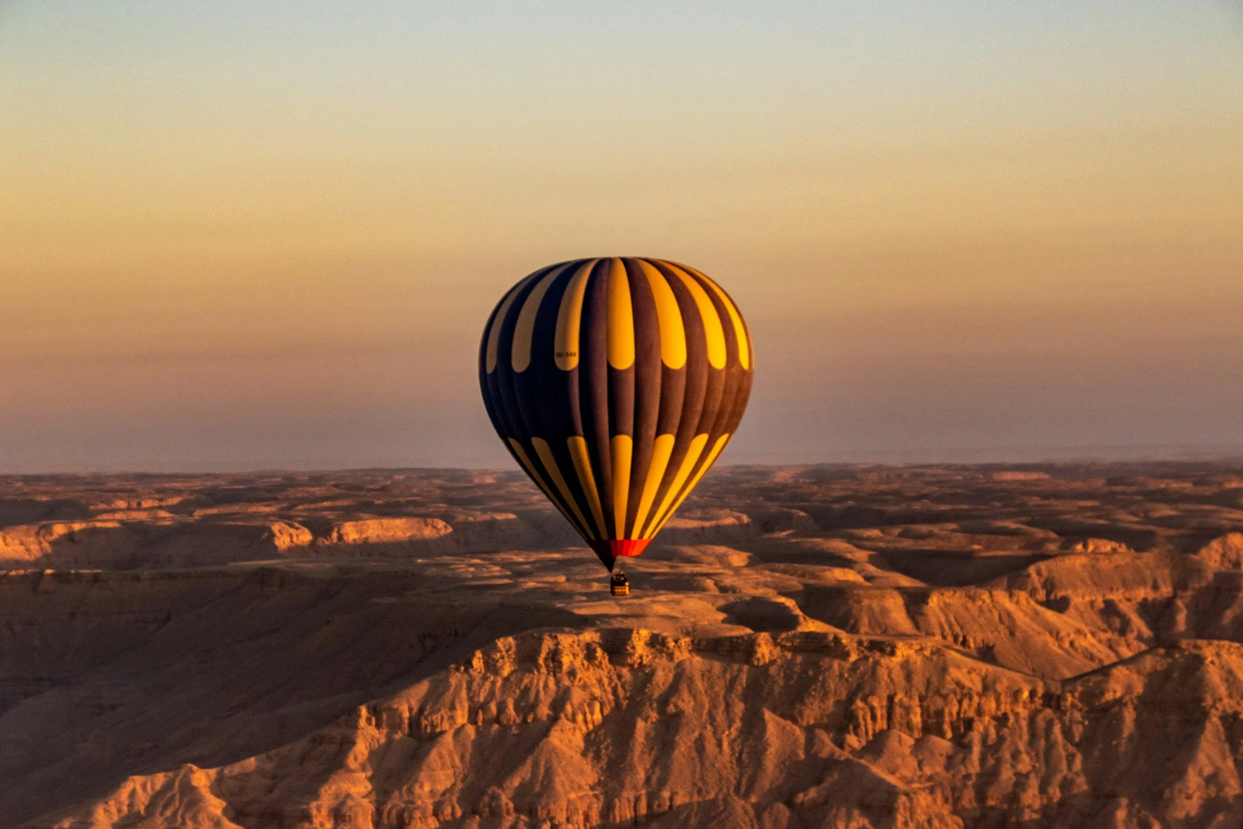 A hot air balloon with black and yellow stripes floating over a desert landscape at sunset.