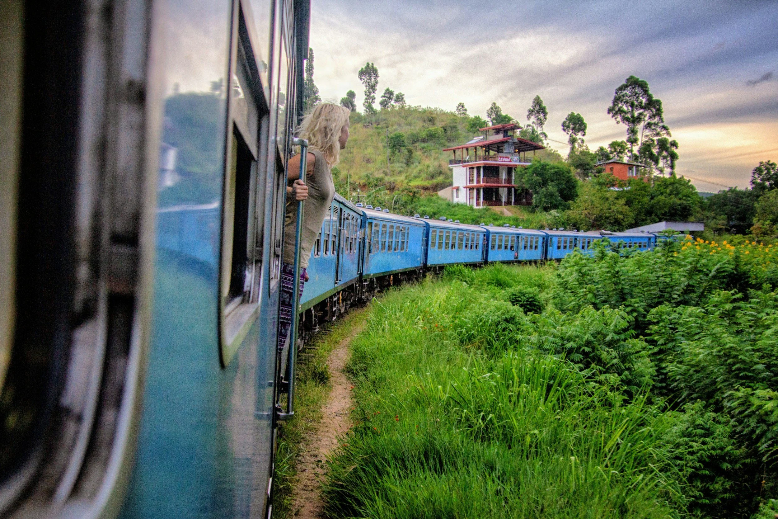 A woman with blonde hair looking out of a blue train window while the train travels through lush green countryside with houses on a hillside during a cloudy sunset.