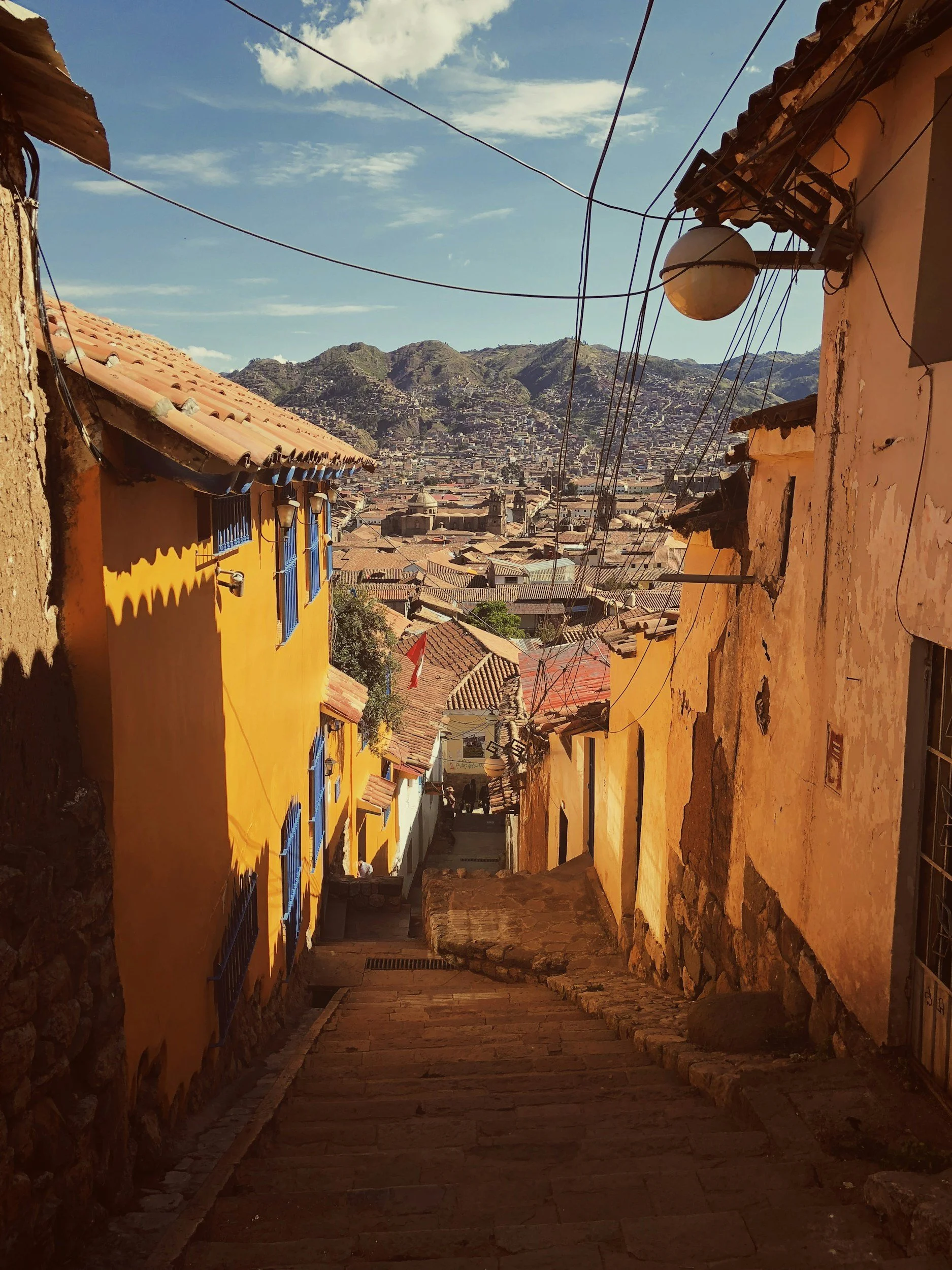 A narrow descending street in a colorful historic neighborhood with yellow and orange buildings, connecting to a scenic view of a city with terracotta rooftops and mountains in the background.