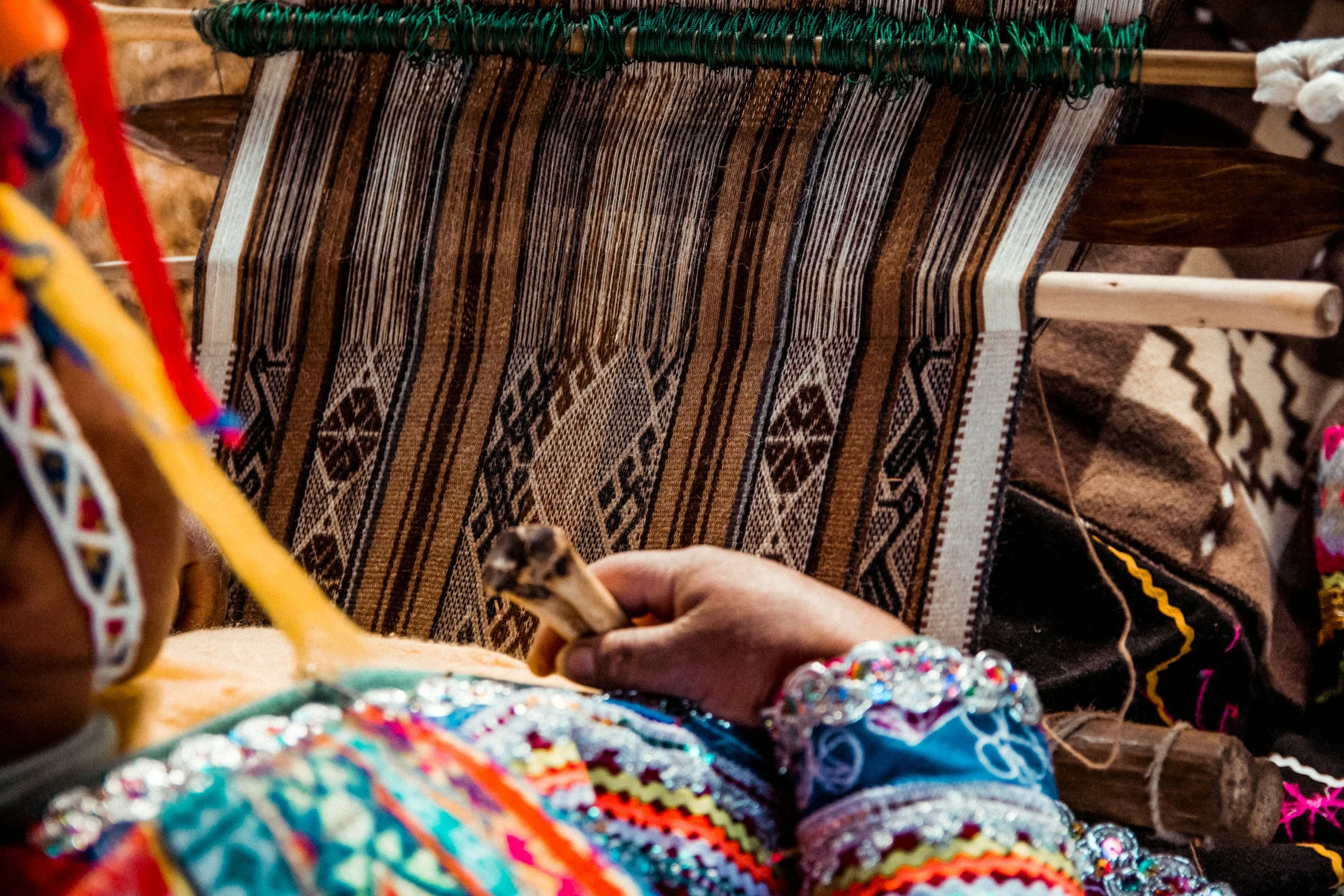 Close-up of hands weaving a traditional patterned textile on a loom, wearing colorful, embroidered clothing.