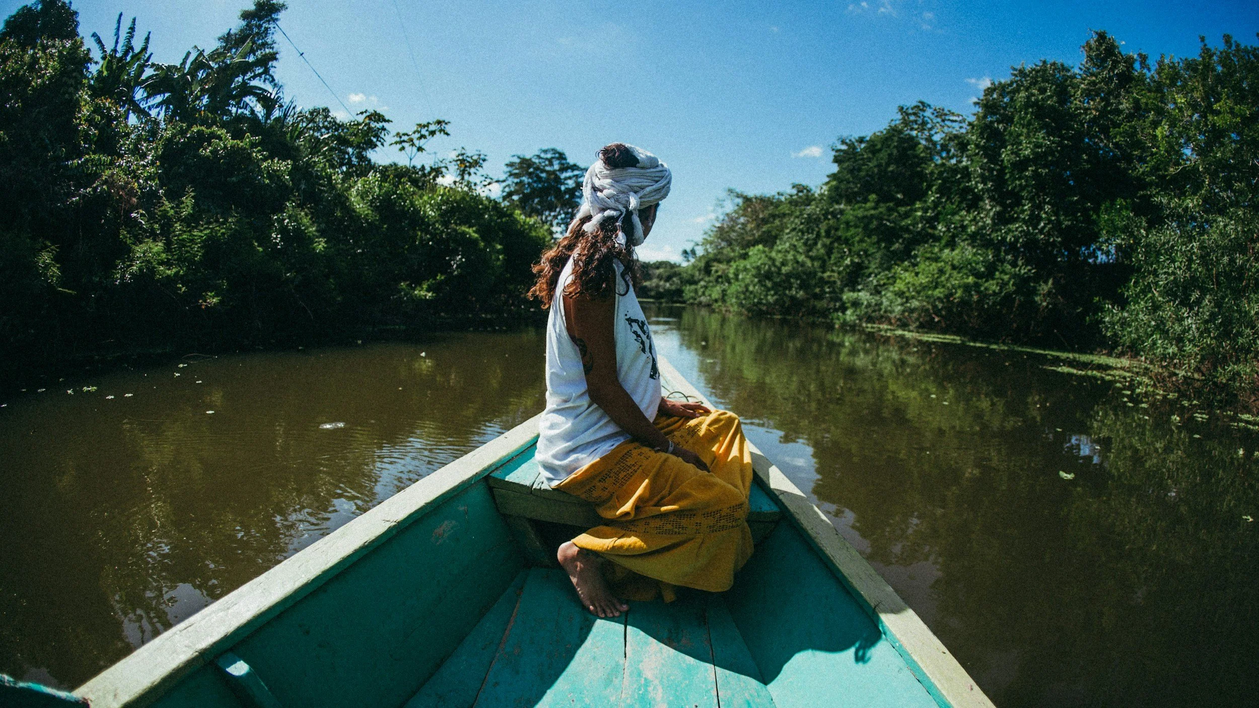 Person sitting on the bow of a boat navigating through a calm waterway surrounded by lush green trees under a bright blue sky.