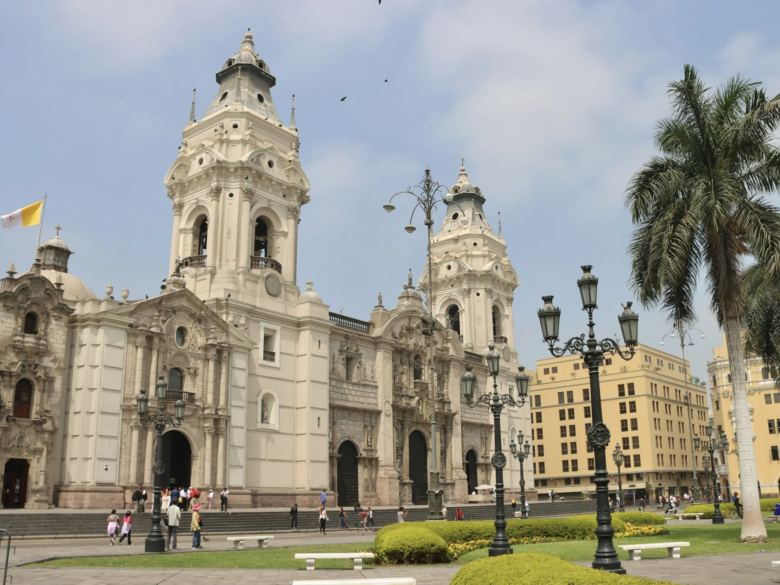 Historic cathedral with two tall towers, surrounded by street lamps, a palm tree, and people walking in the plaza.
