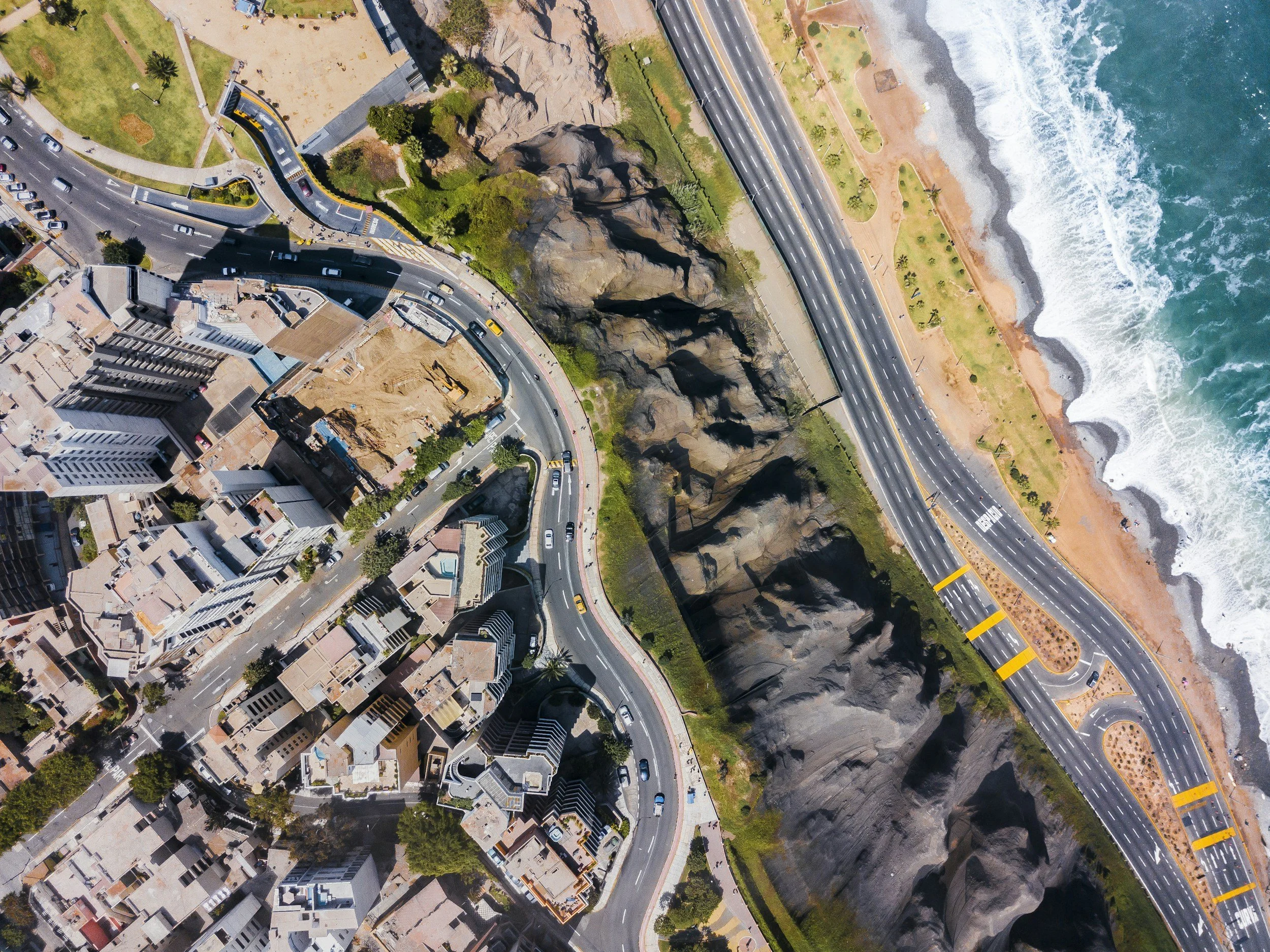 An aerial view of a coastal city with buildings, winding roads, rocky cliffs, a beach, and the ocean waves.