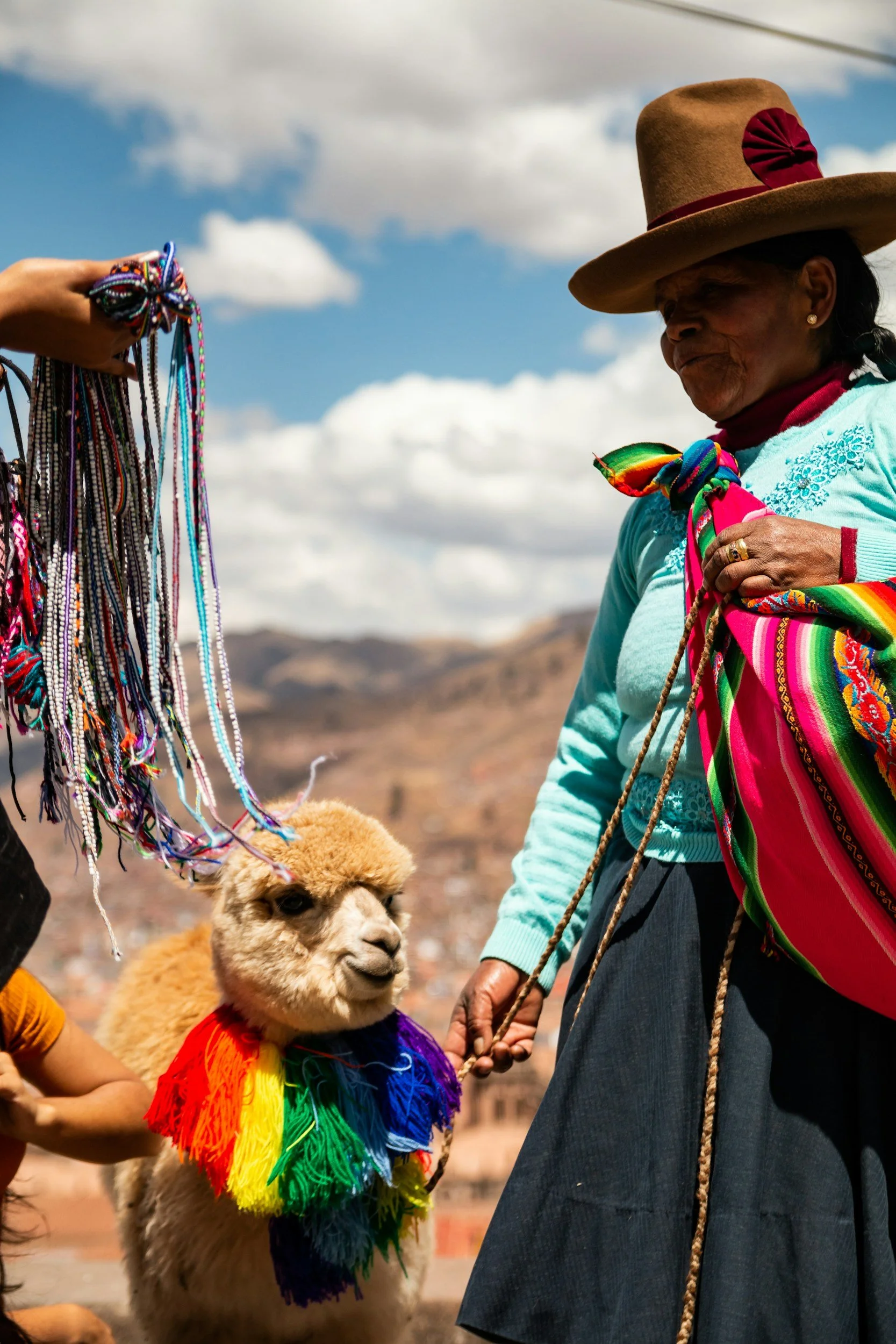 A woman in traditional clothing holding a colorful rainbow scarf and a llama wearing a rainbow-colored fringe with a rainbow scarf around its neck, with another person's hand holding a bundle of colorful beaded necklaces. The background shows a mount