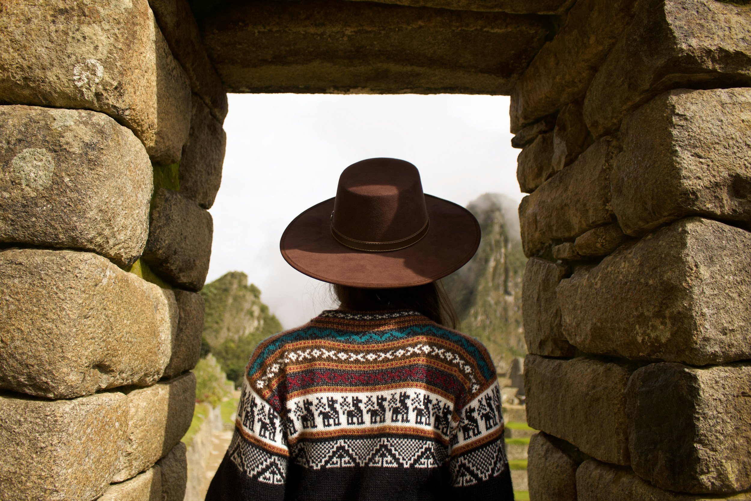 Person wearing a wide-brimmed brown hat and a colorful patterned sweater, standing between stone walls, looking at distant mountains.