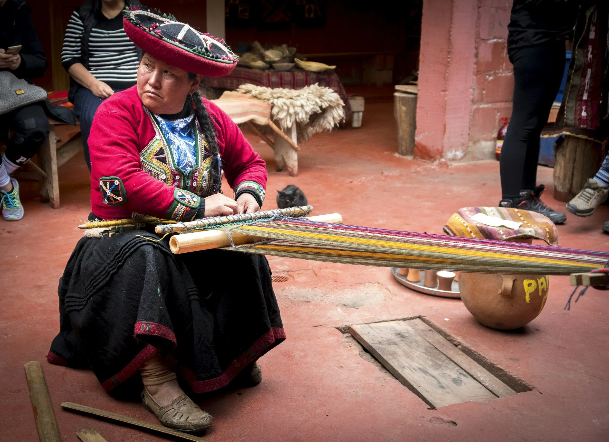A woman dressed in traditional indigenous clothing and a large embroidered hat is weaving on a backstrap loom in a market or workshop setting. Several people are around, some standing and others sitting, with various traditional and modern items visible in the background.