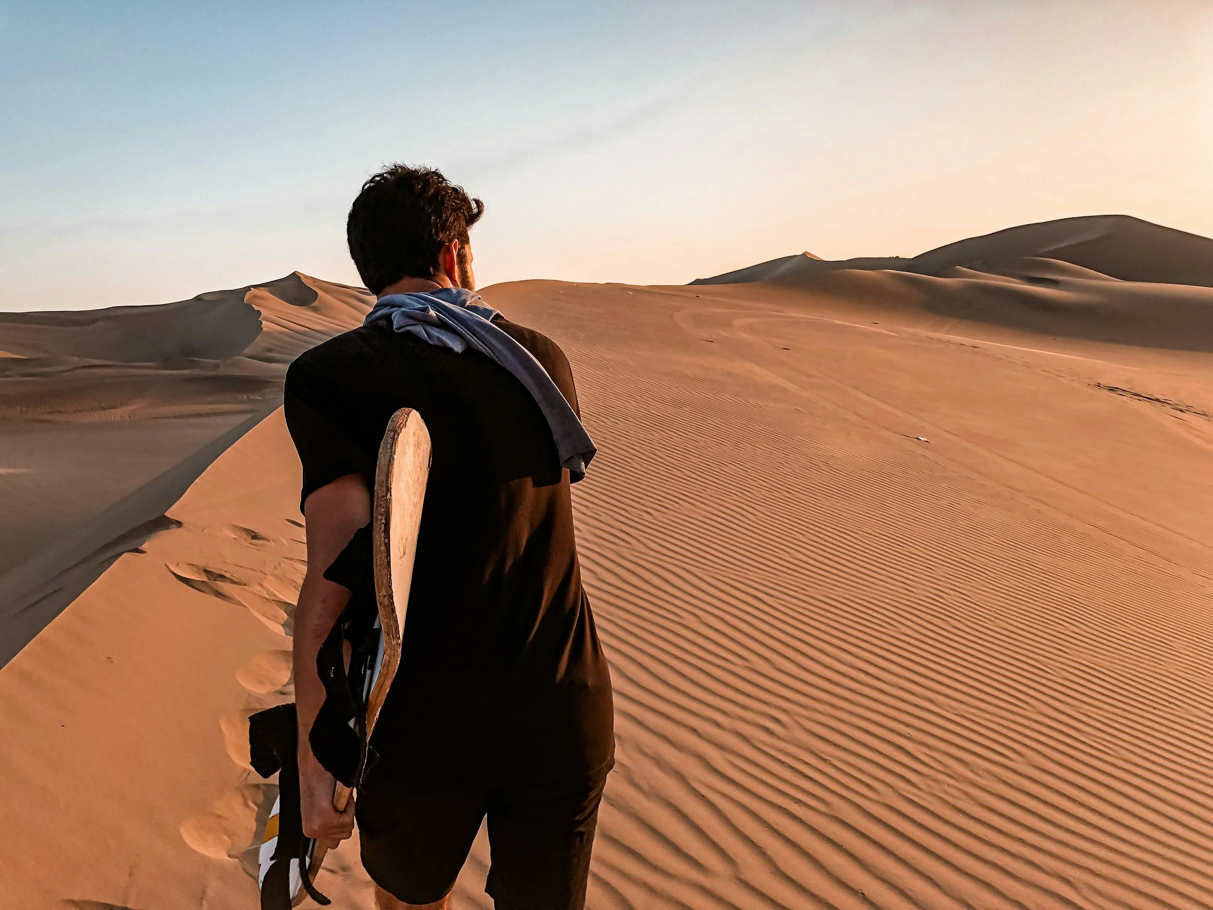 A man walking through a desert with sand dunes, holding a skateboard in his right hand, wearing a black shirt and a shirt draped over his shoulders, during sunset.