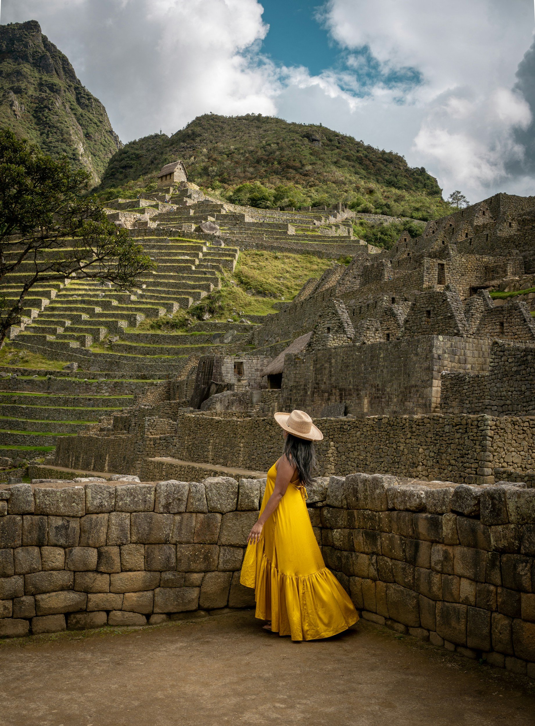 A woman in a yellow dress and wide-brimmed hat standing by a stone wall with ancient Incan ruins and terraced hillside in the background at Machu Picchu.