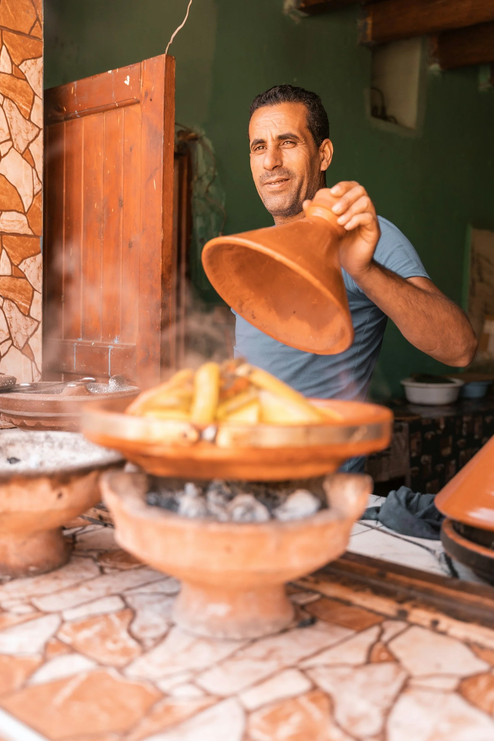 A man is serving food from a traditional clay dish in a rustic kitchen setting with a tiled countertop and stone wall accents.