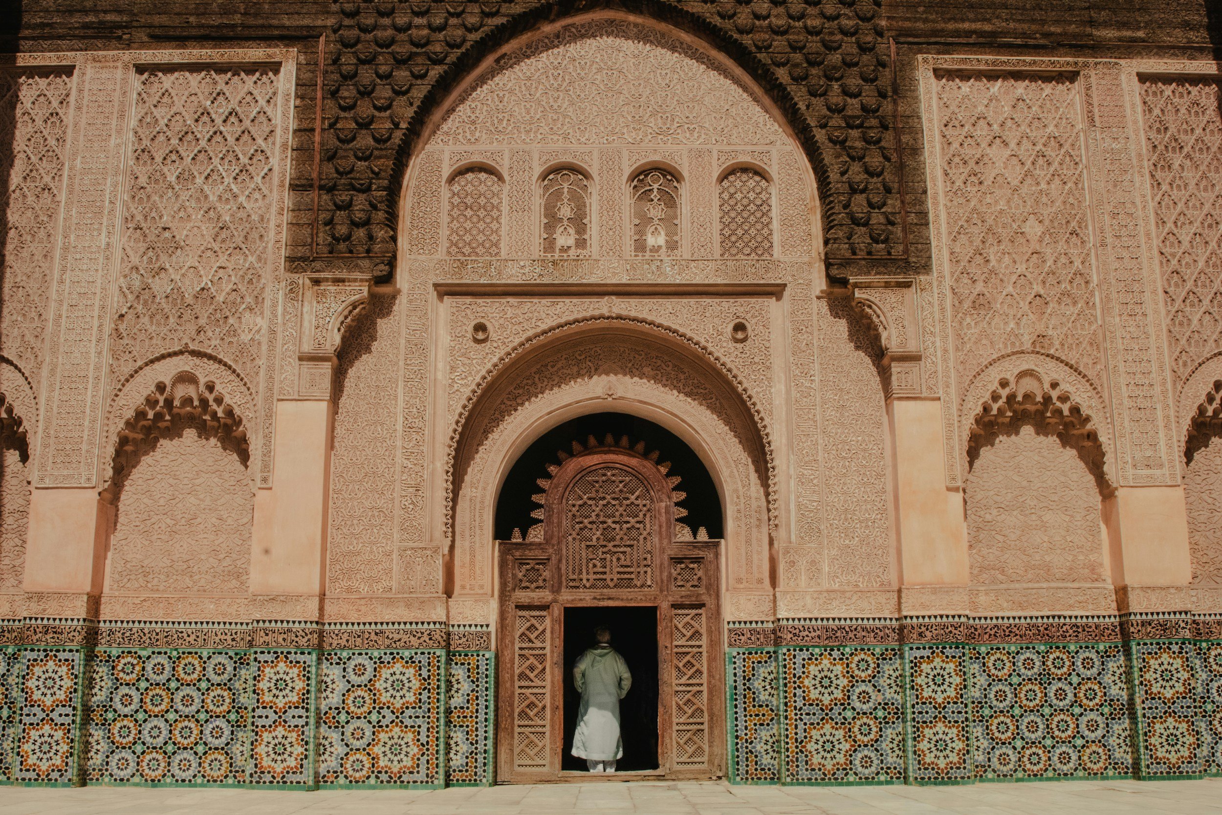 A person in traditional clothing walking into an intricately decorated mosque with carved stonework and colorful mosaic tiles on the lower part of the wall.
