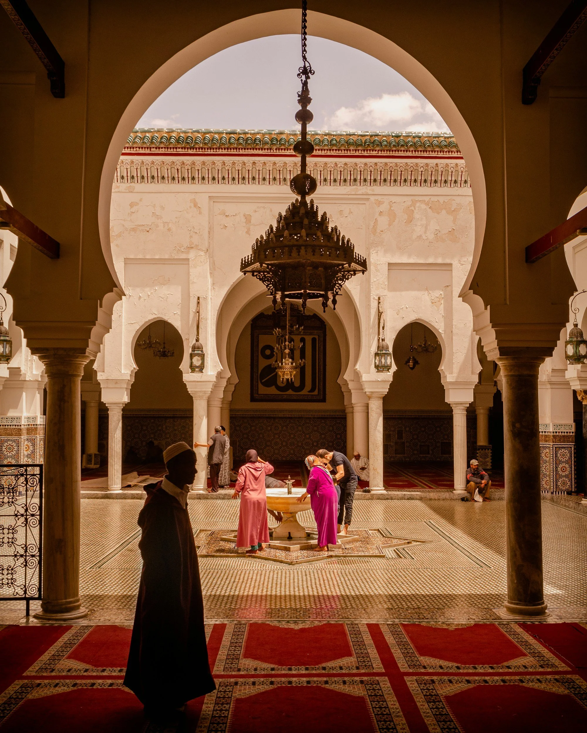 People praying at a fountain inside a mosque with intricate architecture, arches, and hanging lamps.