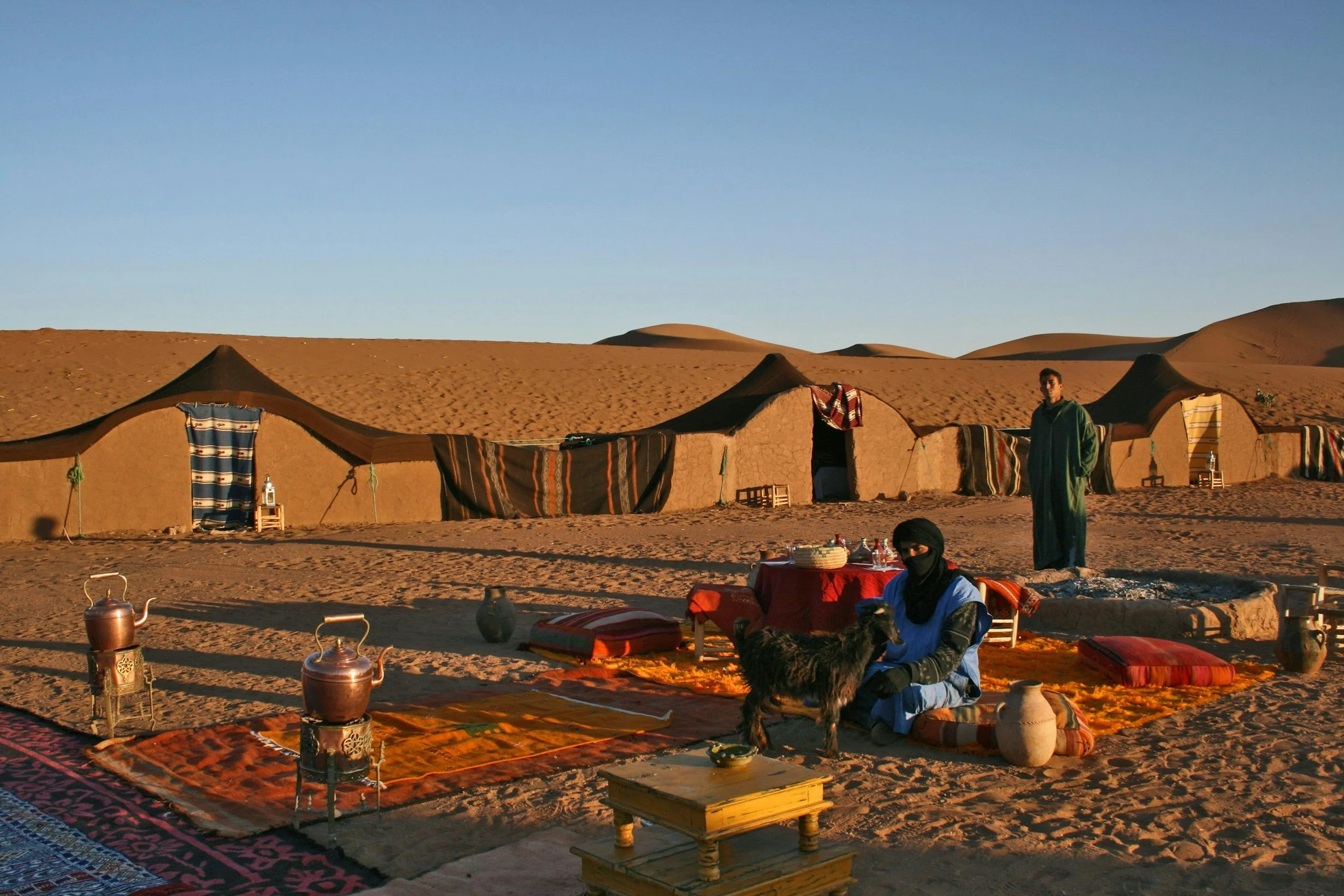 Desert campsite with traditional tents, a woman in a blue dress and black headscarf sitting with a dog, man in green robe standing nearby, and desert dunes in the background.