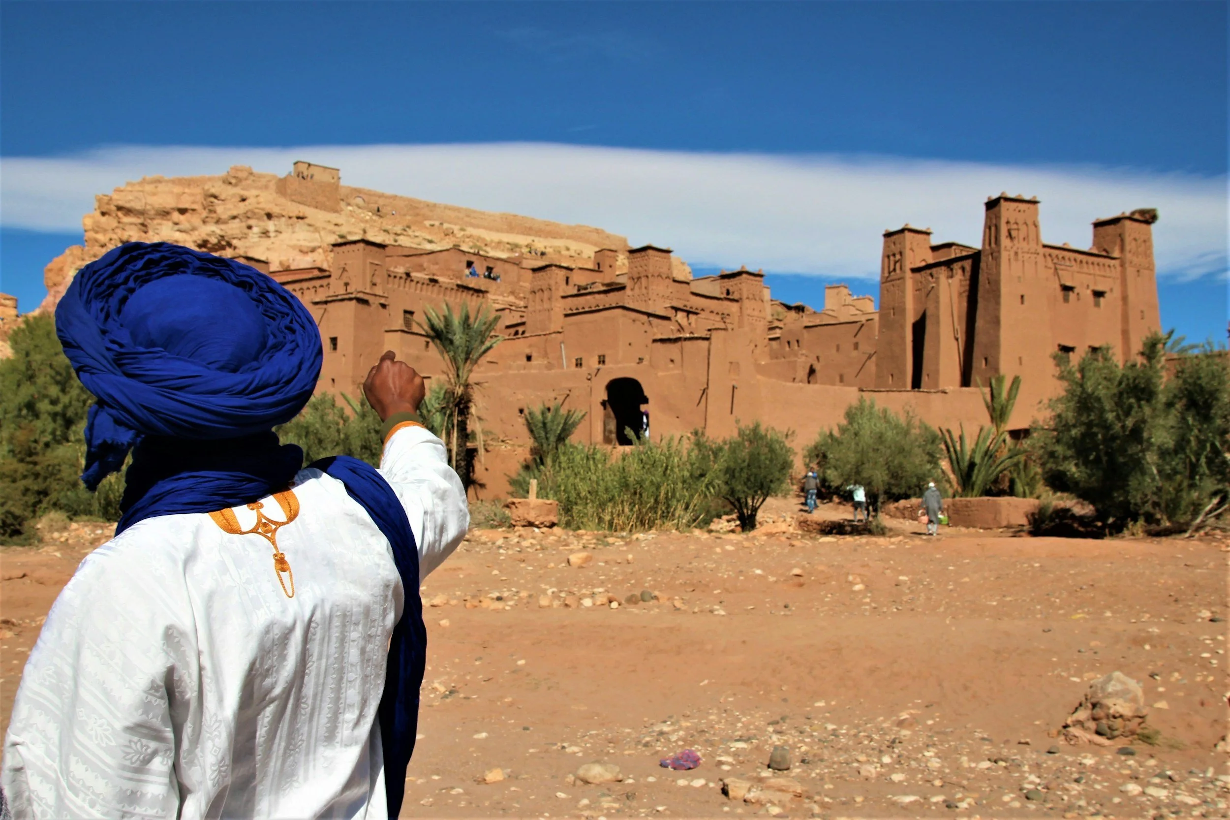 A person wearing a white traditional djellaba and a large blue turban, facing a historic mud-brick fortified village on a desert landscape with sparse vegetation, under a blue sky with clouds.
