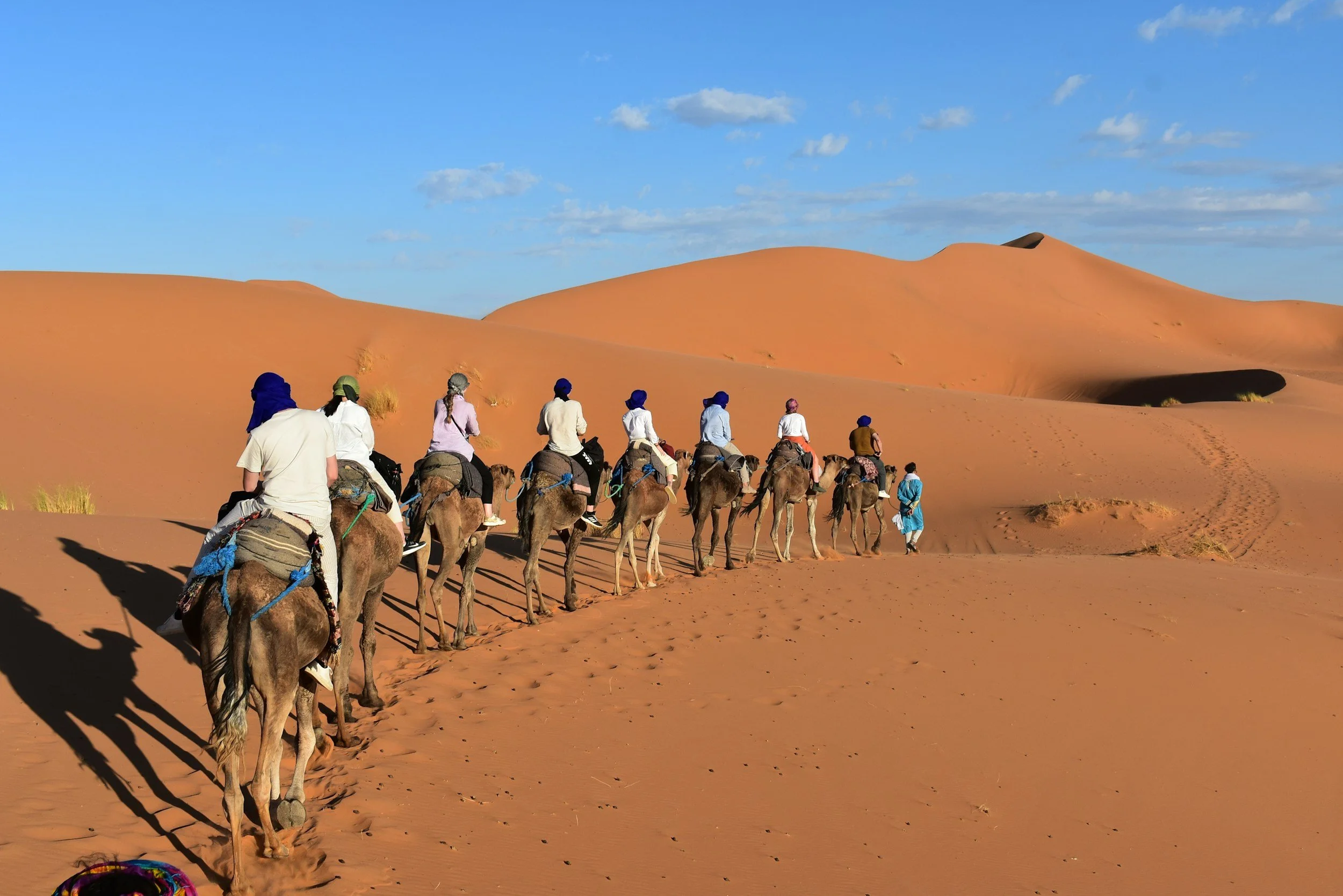 A group of tourists riding camels through a desert landscape with sand dunes under a blue sky with scattered clouds.