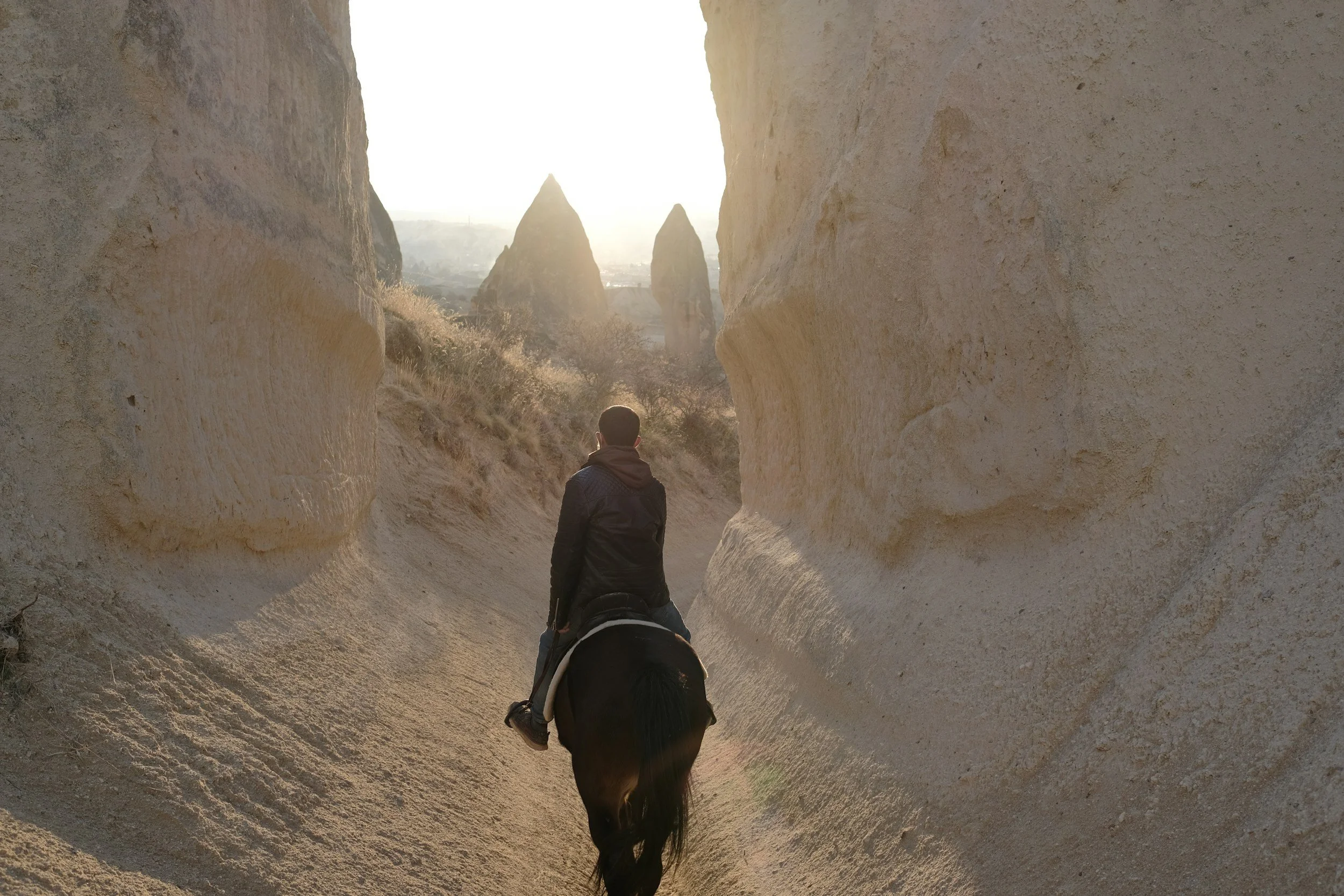 A person riding a horse through a narrow, rocky canyon with large, beige rock formations on either side and a bright sun in the distance.