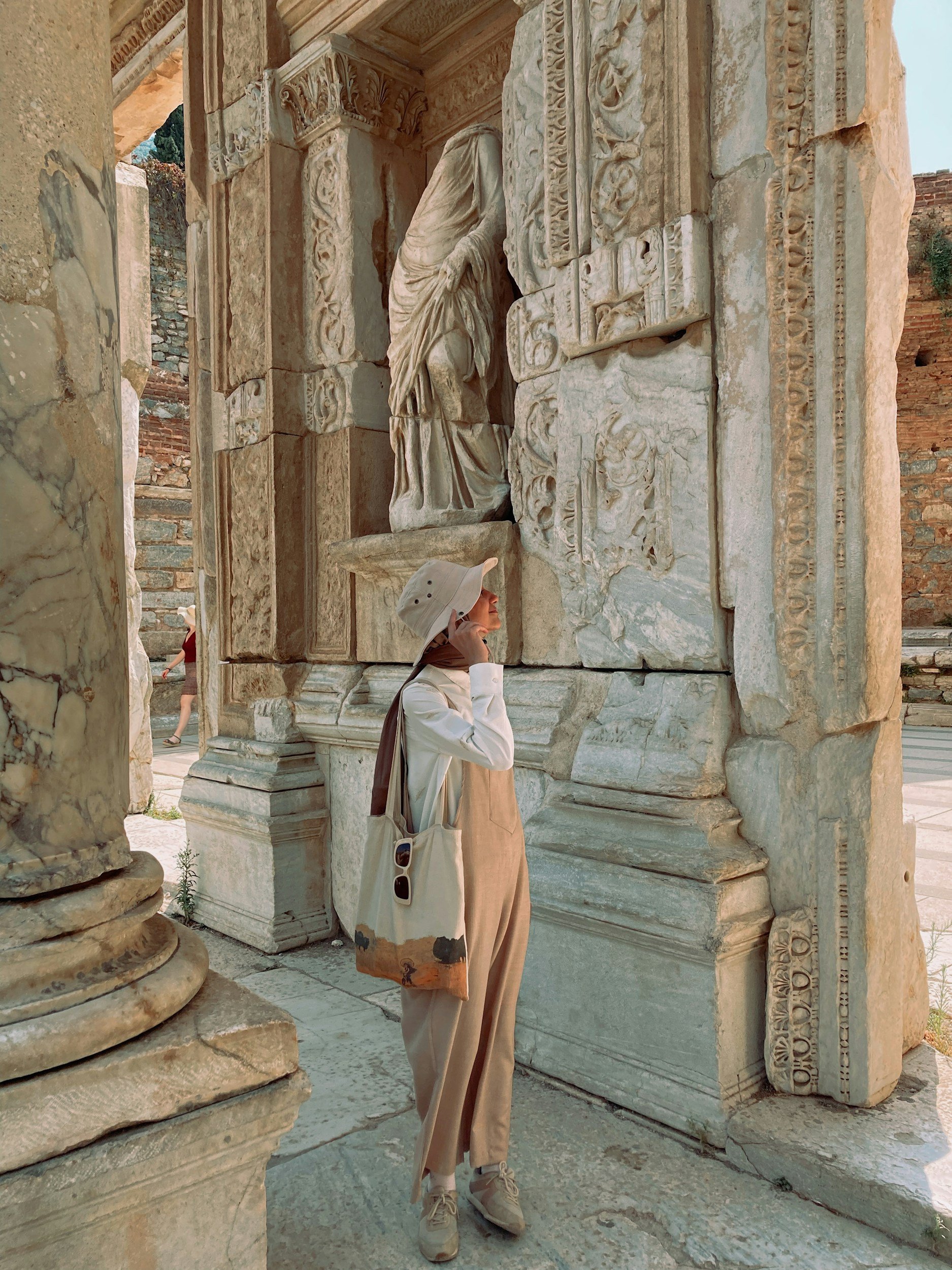 A woman in beige clothing, hat, and tote bag stands next to ancient stone ruins with intricate carvings and statues, possibly part of an archaeological site.
