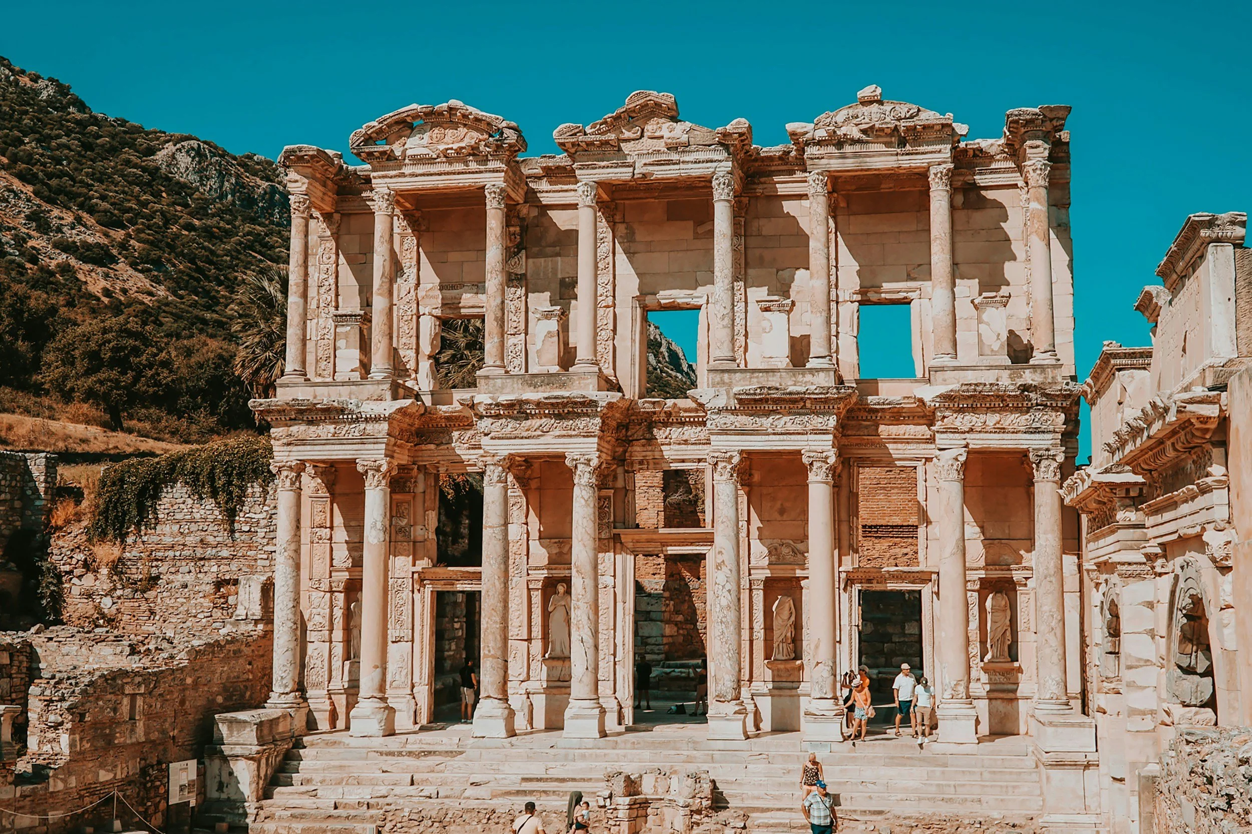 Ancient Roman theater facade with columns and statues, surrounded by tourists, against a backdrop of a mountain and clear blue sky.