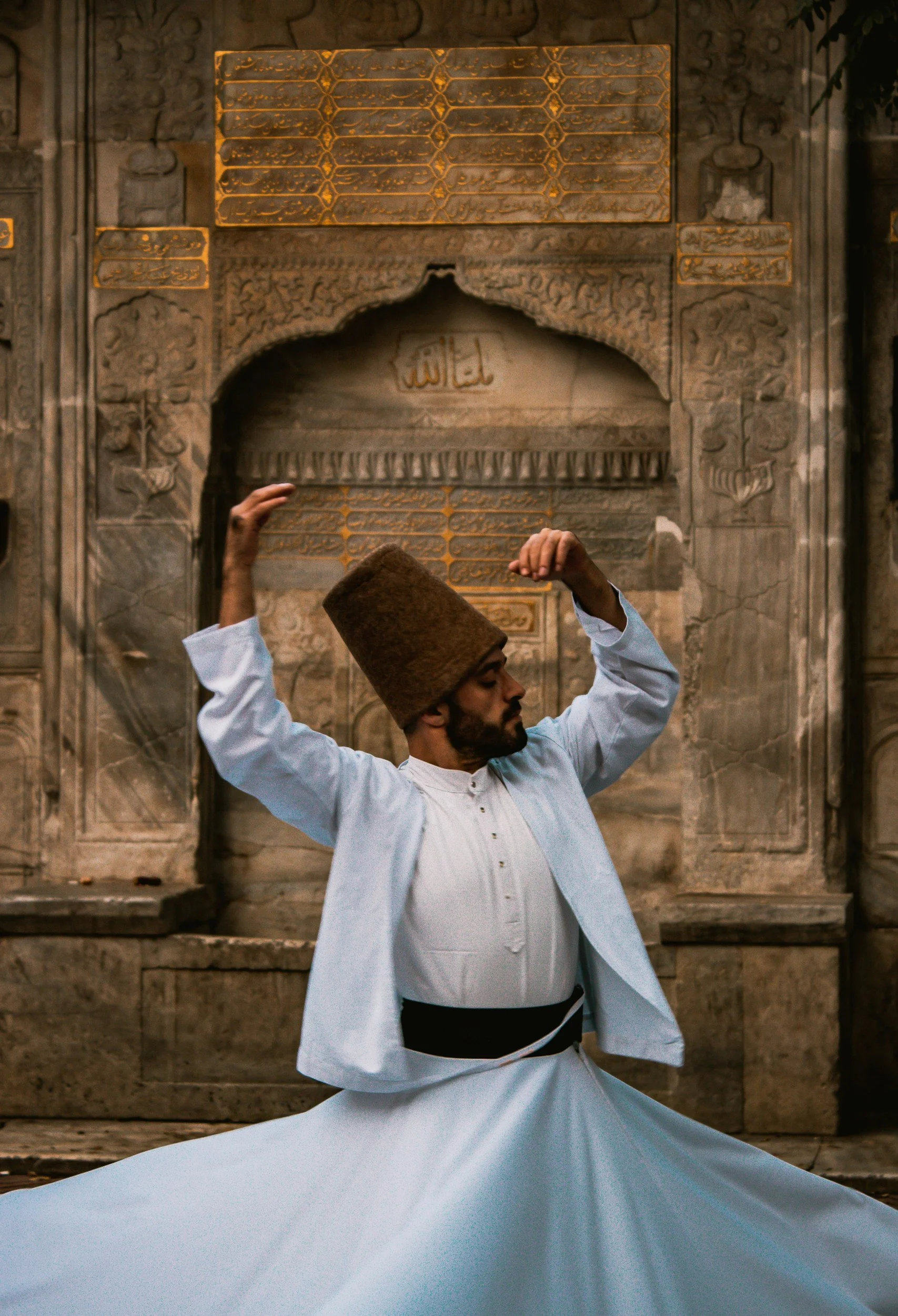 A man wearing traditional apparel, including a tall brown hat and a white robe, is performing a dance in front of an ornate, carved stone wall with Arabic script.