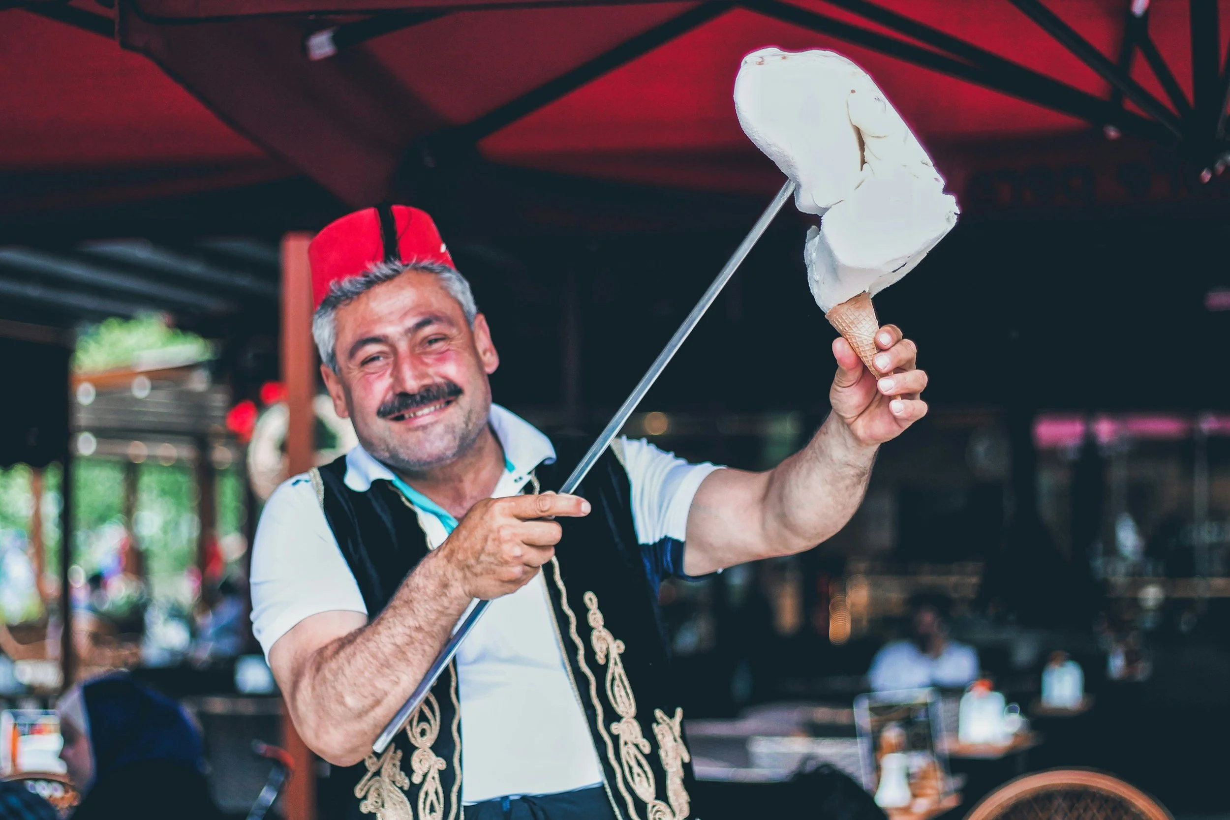 A man with a mustache smiling, wearing a red fez hat and a traditional vest, holding a large ice cream cone topped with whipped cream, and a rod with a scoop of ice cream, in an outdoor setting.