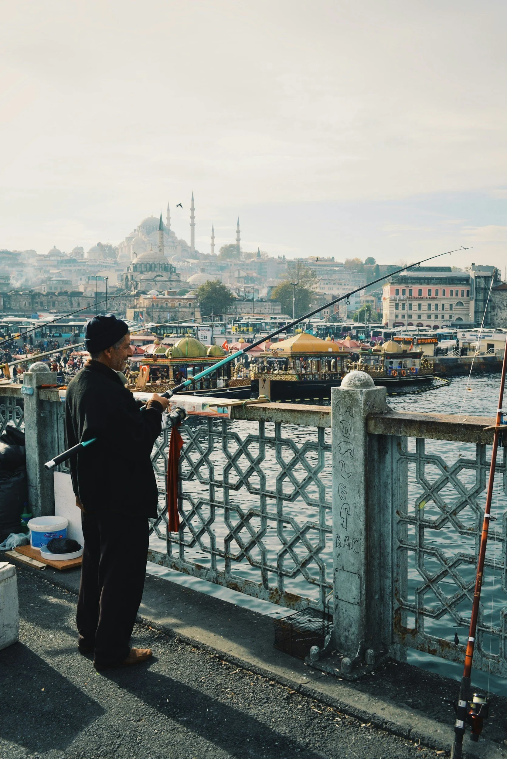 A man fishing by a river with a view of a historic cityscape, including mosques with minarets, in the background.