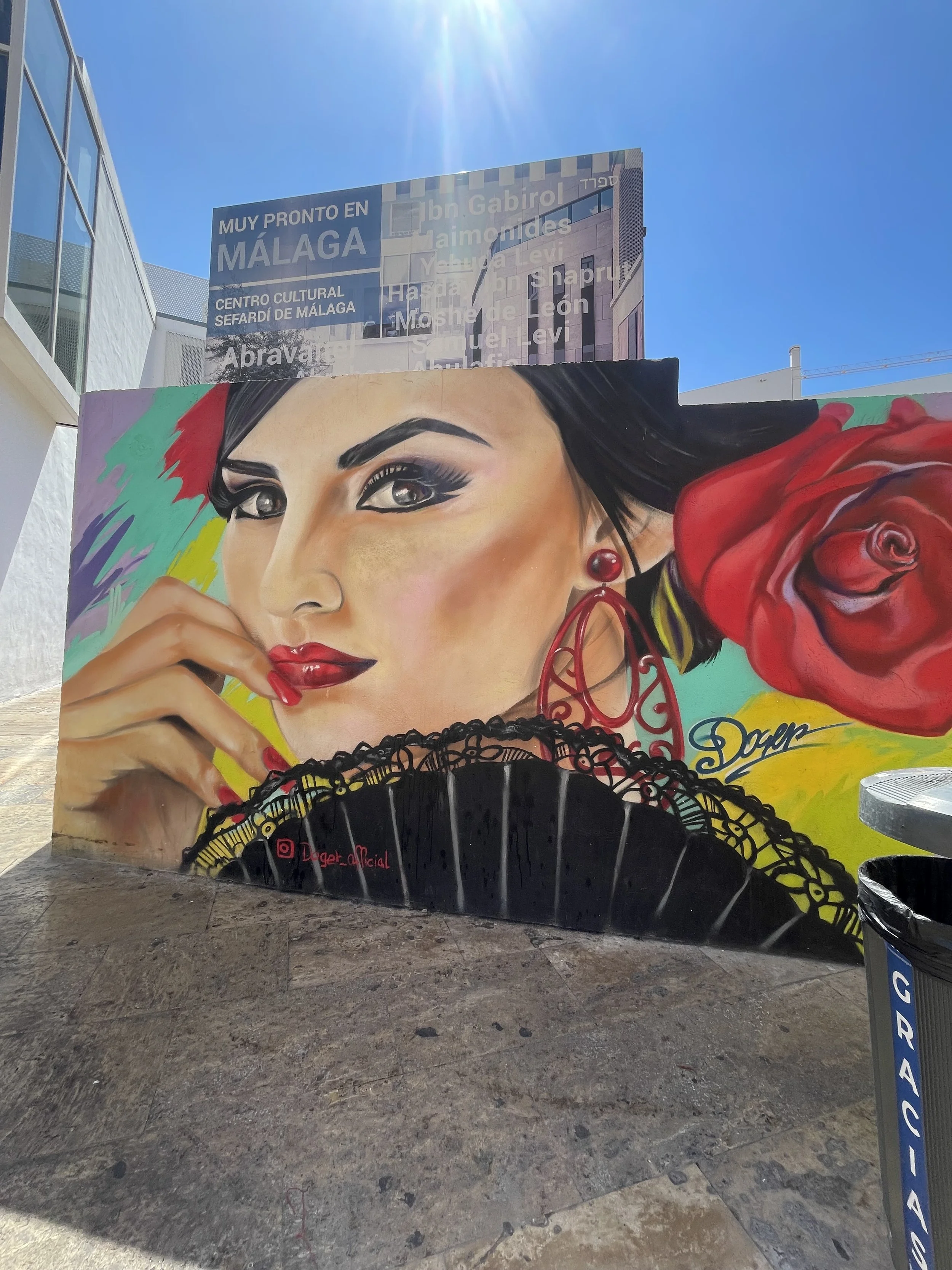 Colorful mural of a woman with dark hair, red lipstick, and earrings, holding a fan with lacy edges and a red rose in her hair, set against a bright background.