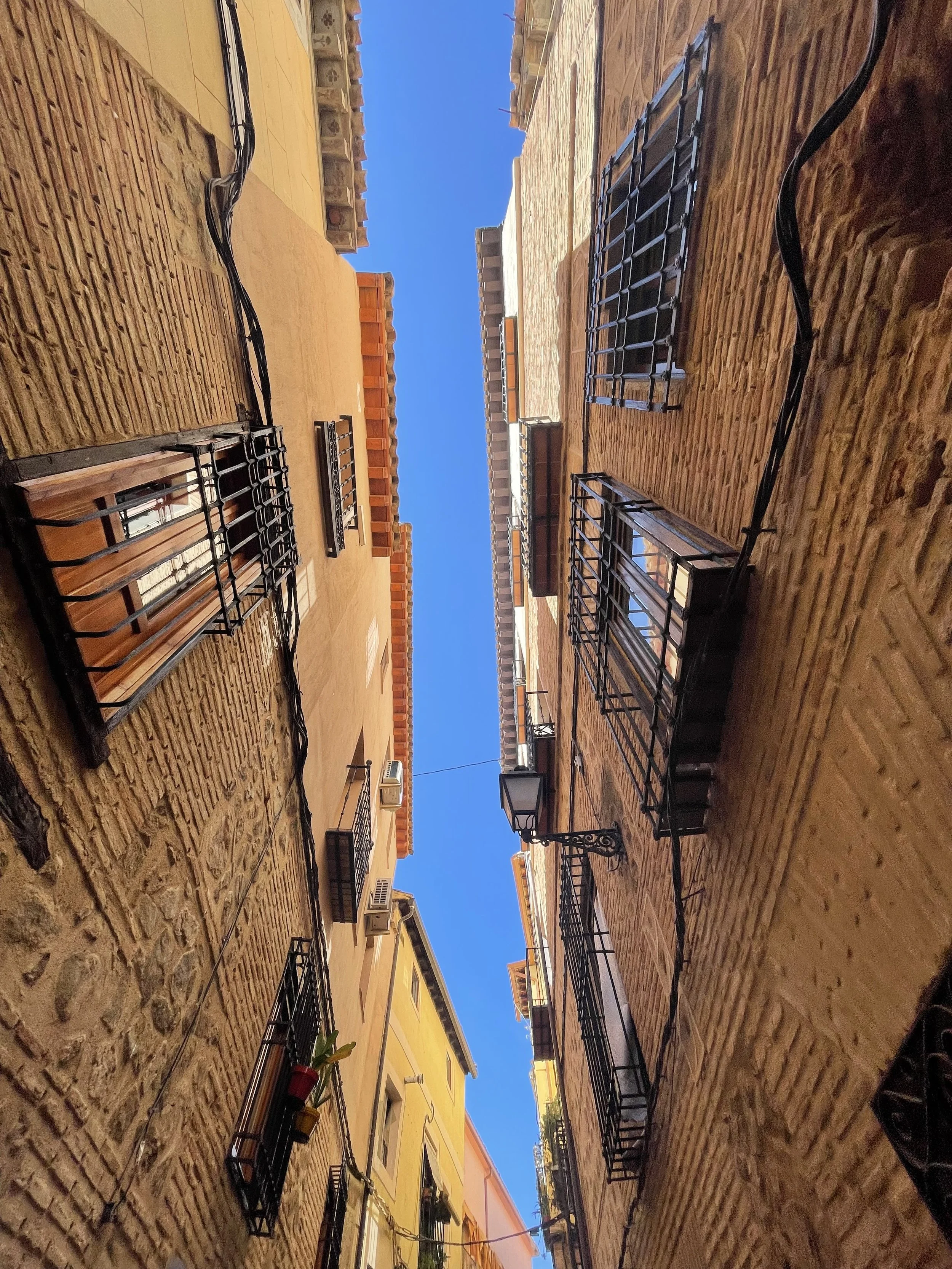 Narrow alleyway between tall brick buildings with black metal balconies, windows, and air conditioning units, under a bright blue sky.