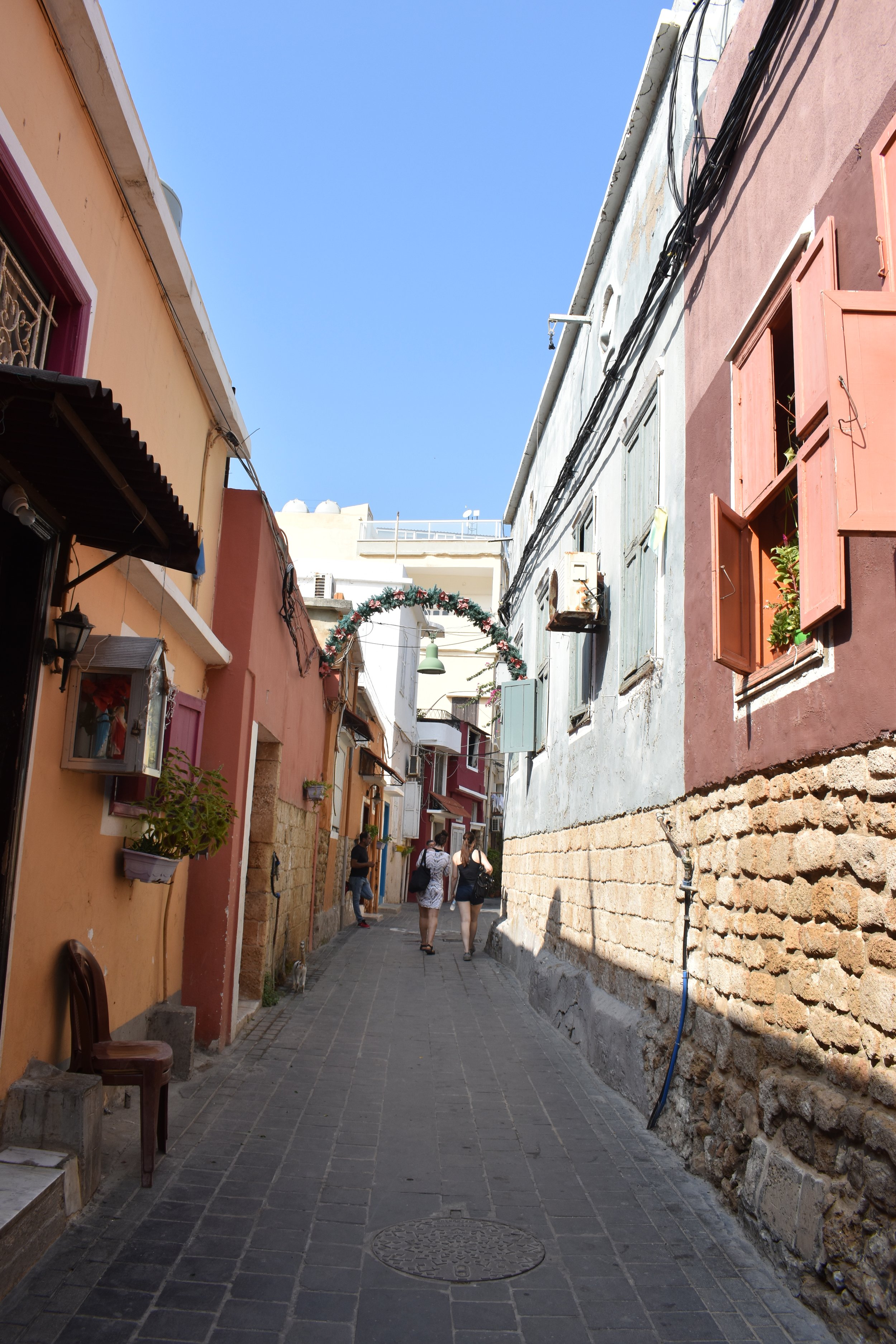 A narrow street in a colorful old town, with pastel buildings, open window shutters, and people walking under a holiday decoration arch.