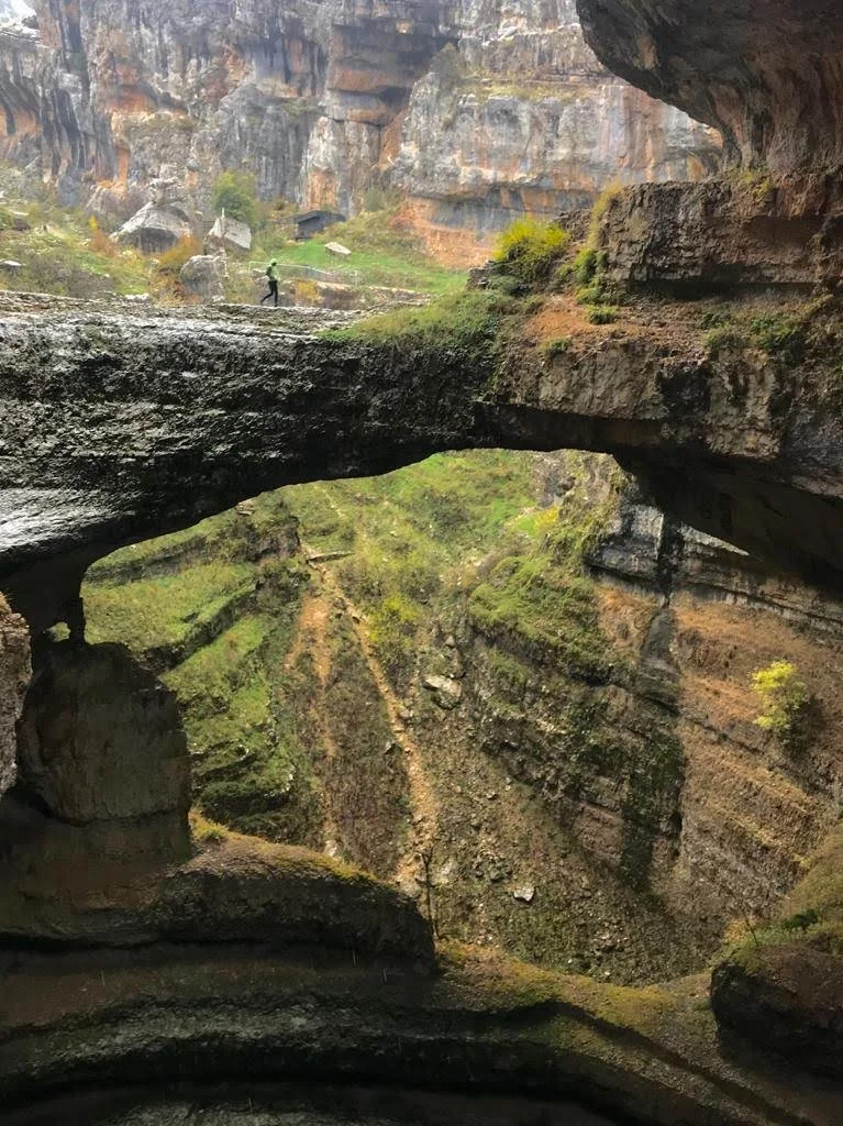 A person hiking on a narrow rock ledge inside a deep canyon with high, layered cliffs and sparse vegetation.