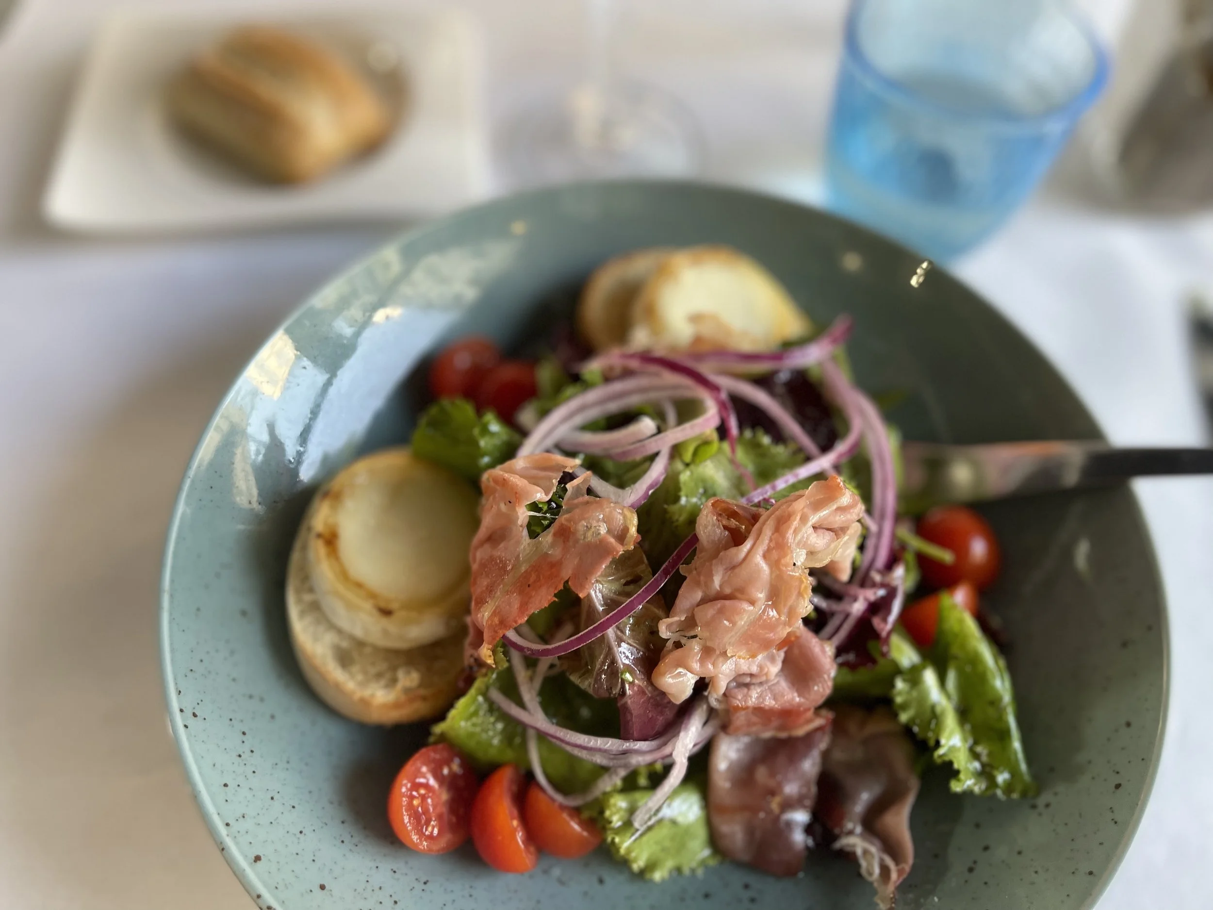 A salad with cherry tomatoes, sliced red onions, hard-boiled eggs, lettuce, and slices of prosciutto in a blue bowl, with a plate of bread and a glass of water in the background.