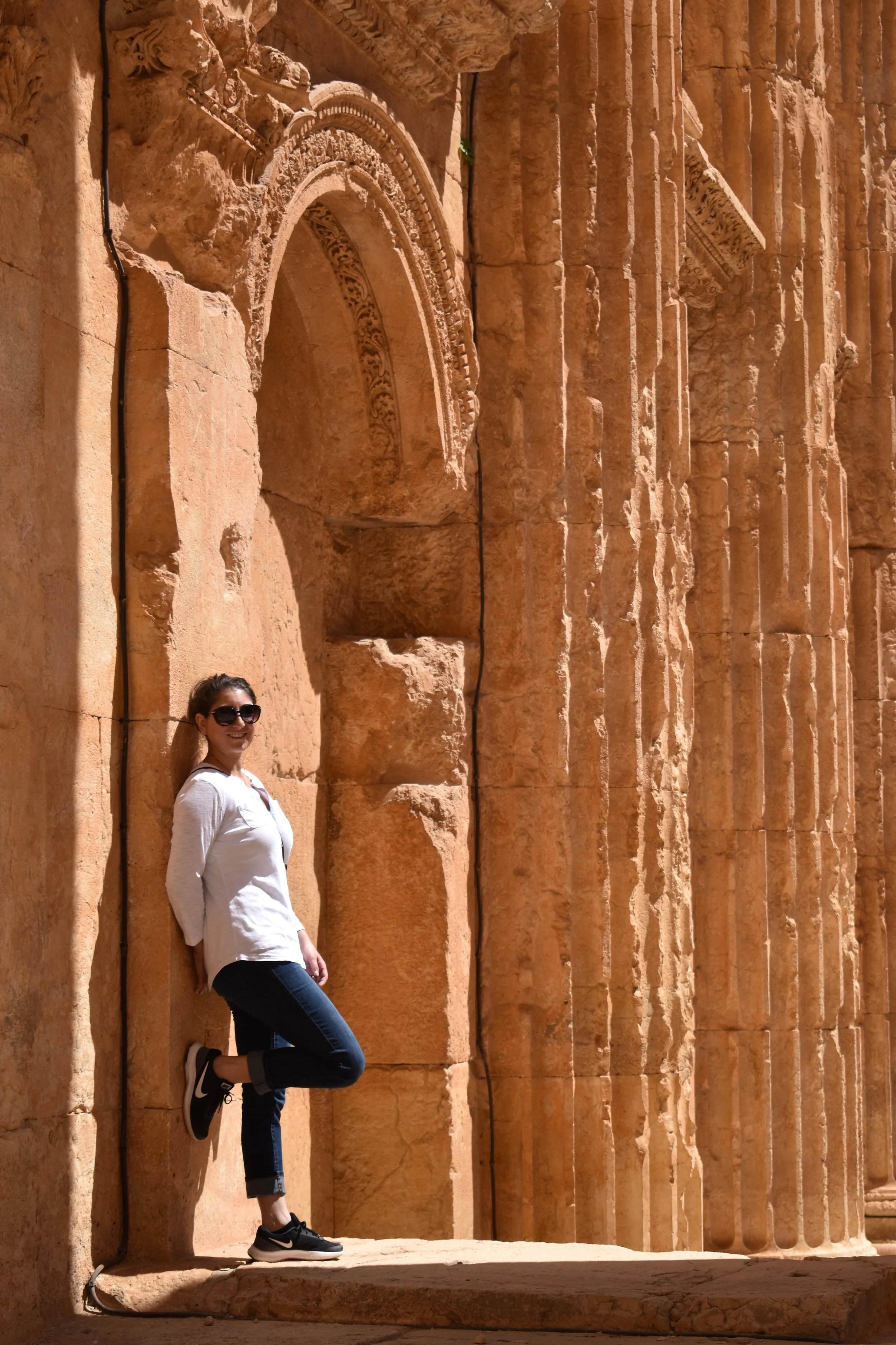 A woman in white shirt and sunglasses leaning against ancient sandstone columns with intricate carvings in an archaeological site.