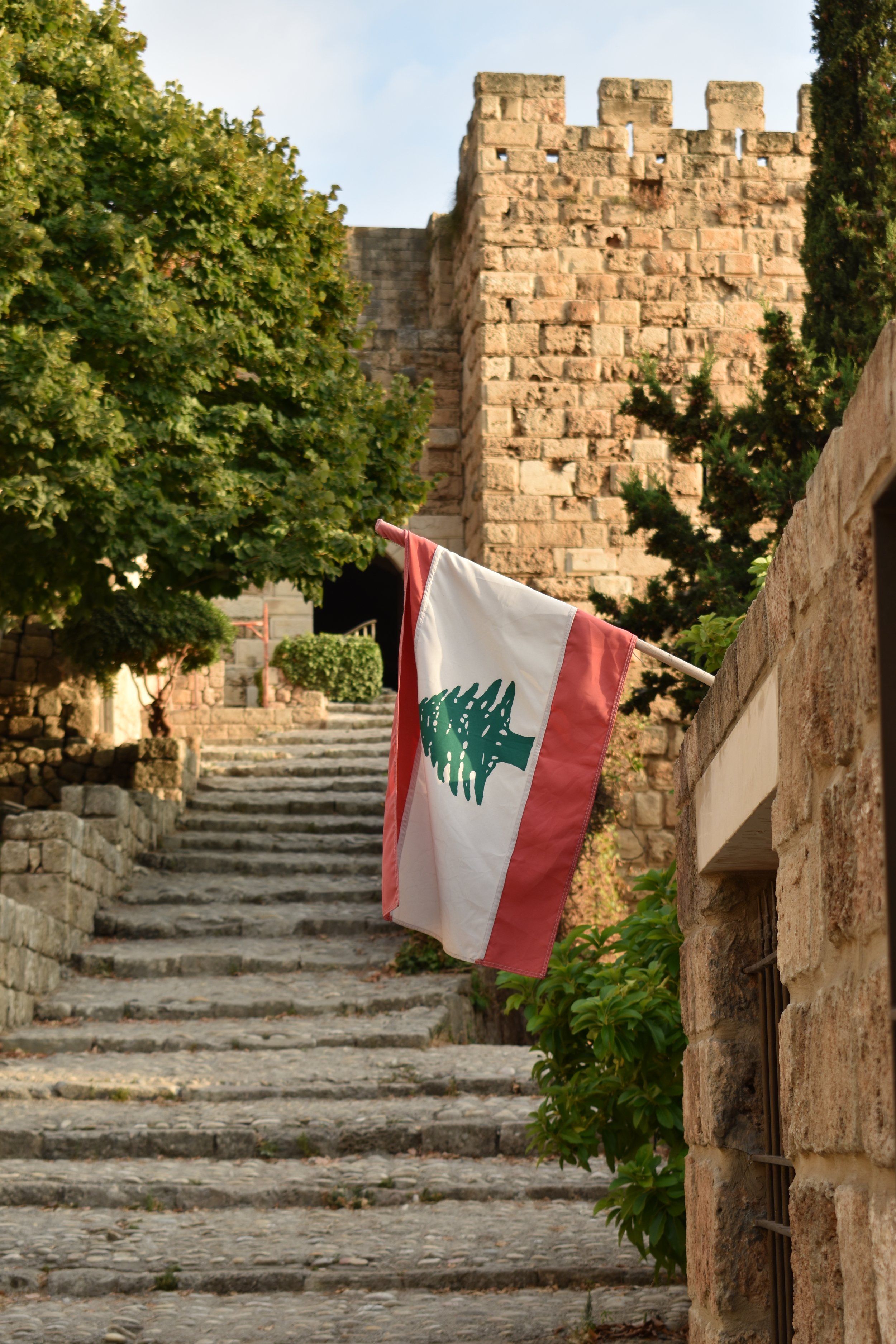 A Lebanese flag hanging by a stone wall with stone steps and a castle tower in the background, surrounded by trees.