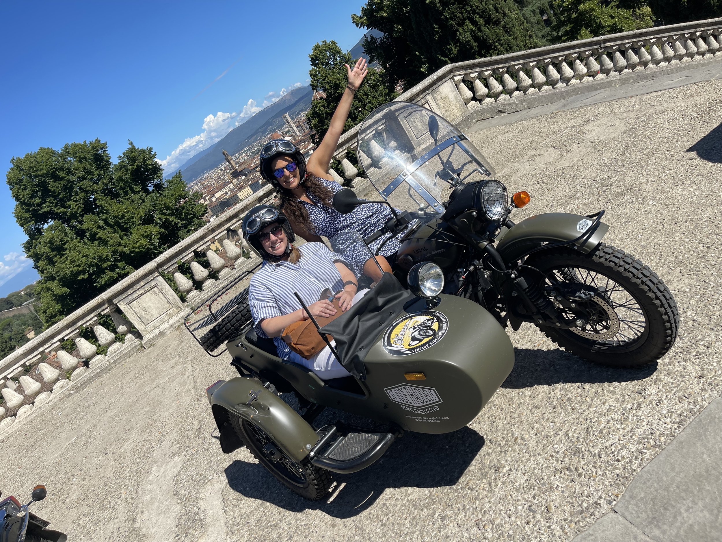Two women riding a vintage motorcycle with a sidecar on a scenic overlook with a cityscape and mountains in the background. Both women are wearing helmets and sunglasses; one is waving.