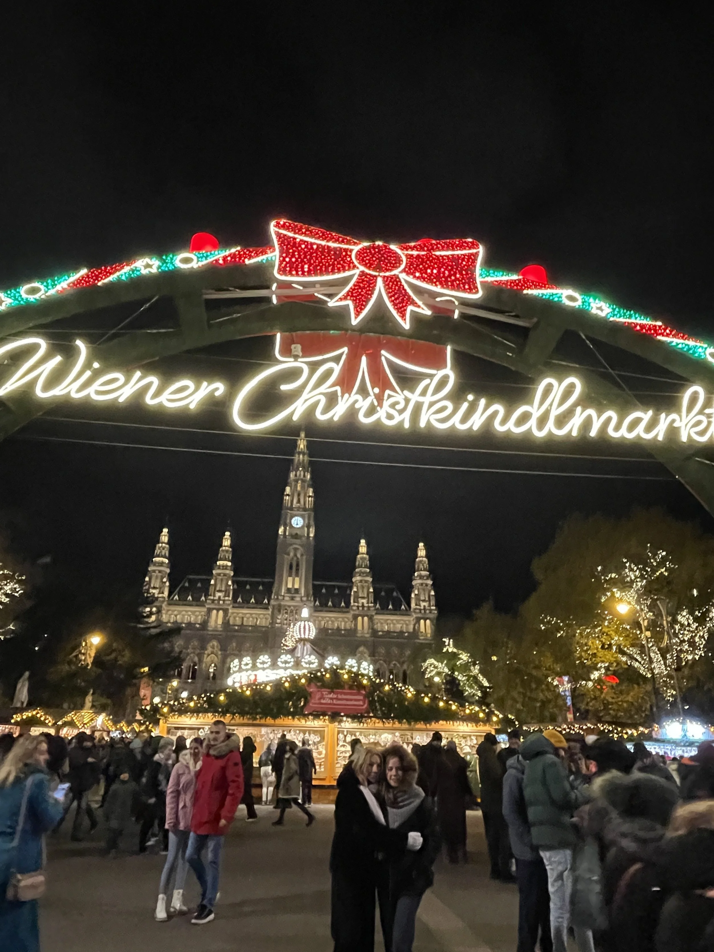 Night scene at Wiener Christkindlmarkt, a Christmas market in Vienna, Austria, with festive lights, a decorated archway, and a historic building in the background.