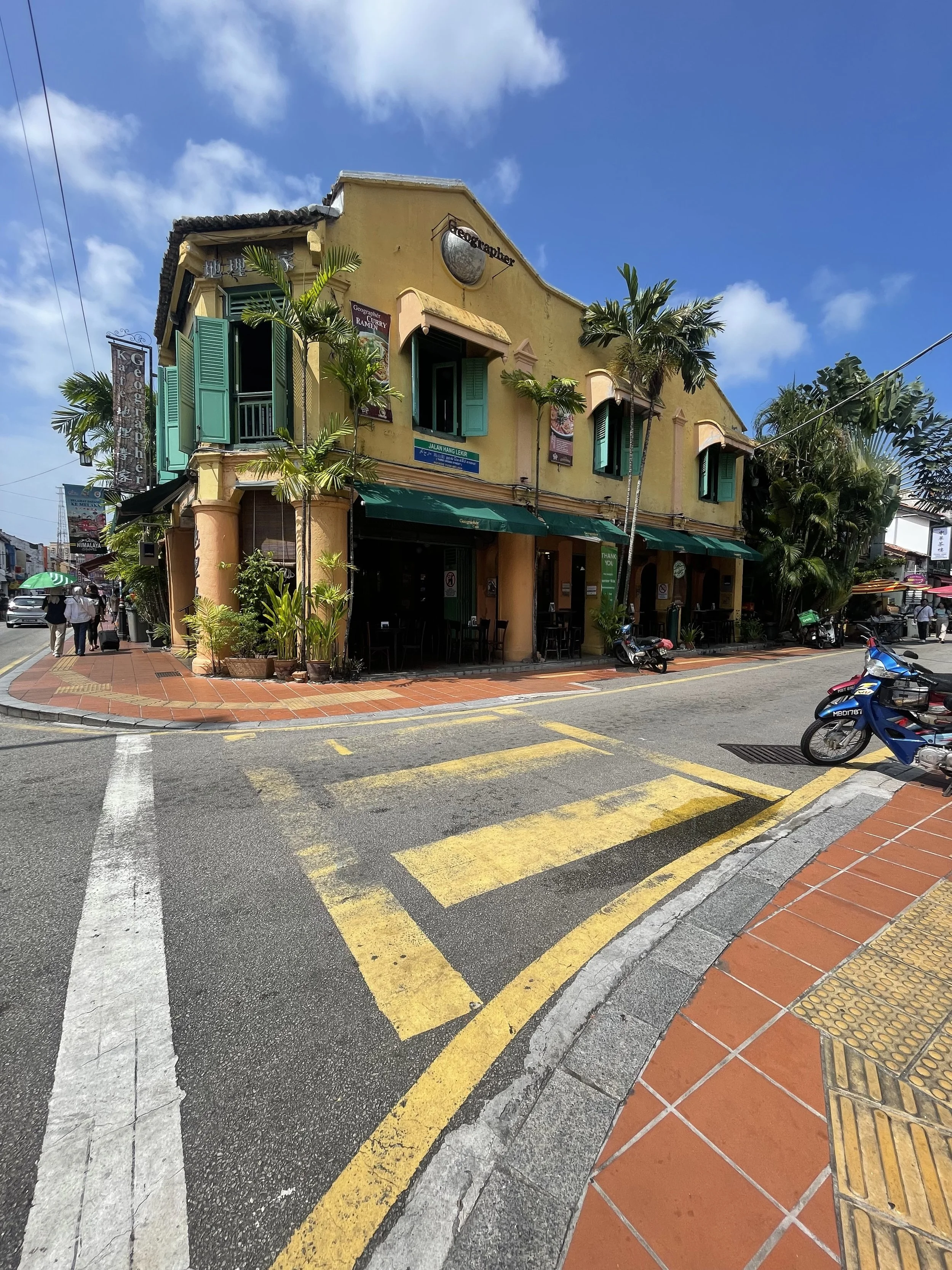 Yellow two-story building with green shutters, palm trees, outdoor seating, and motorcycles parked nearby on a sunny street with a crosswalk