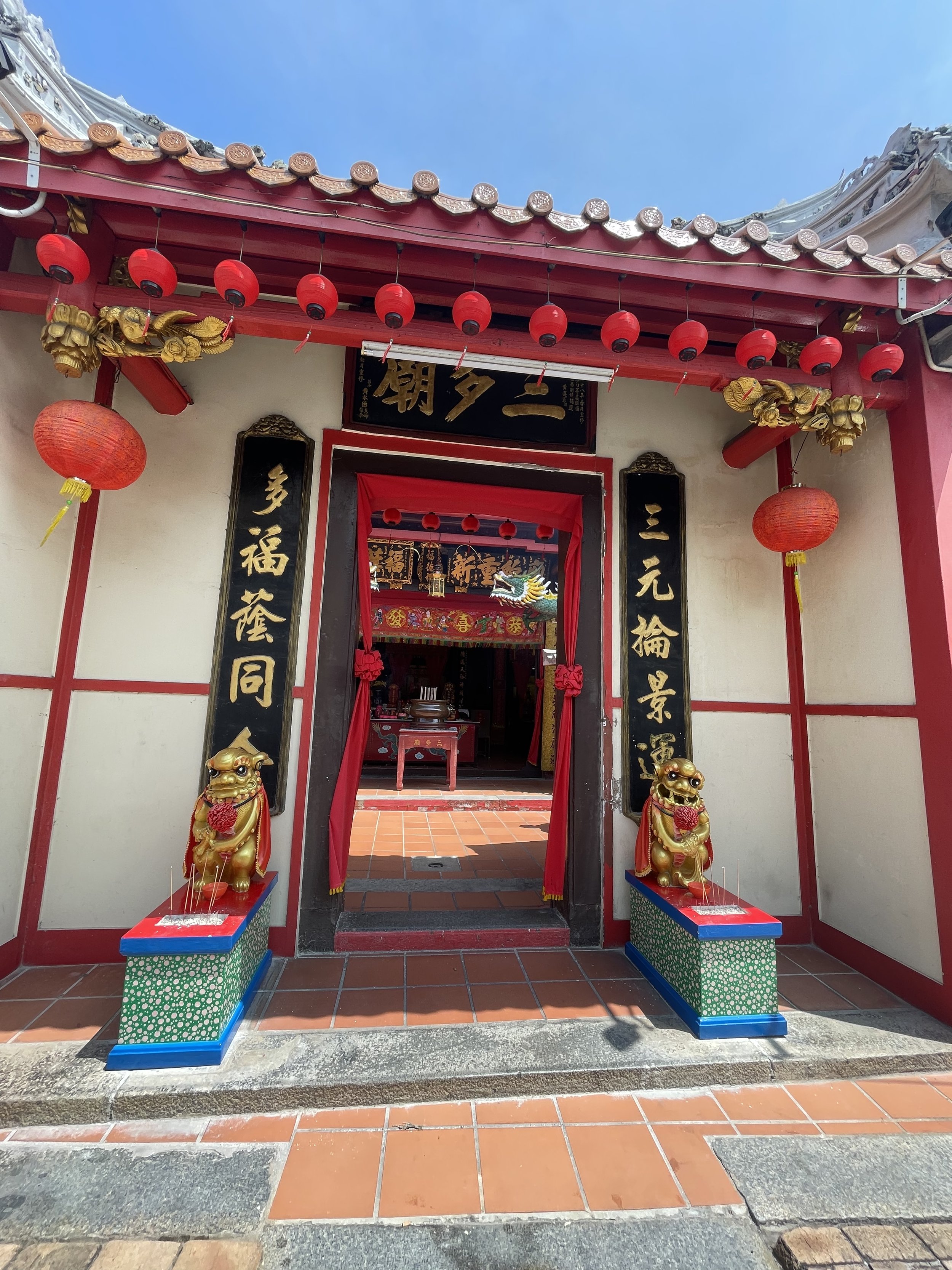 The entrance to a traditional Chinese temple features red lanterns, two golden lion statues, and black plaques with gold Chinese characters. The doorway has red curtains, leading inside to the temple interior decorated with red and gold ornaments.