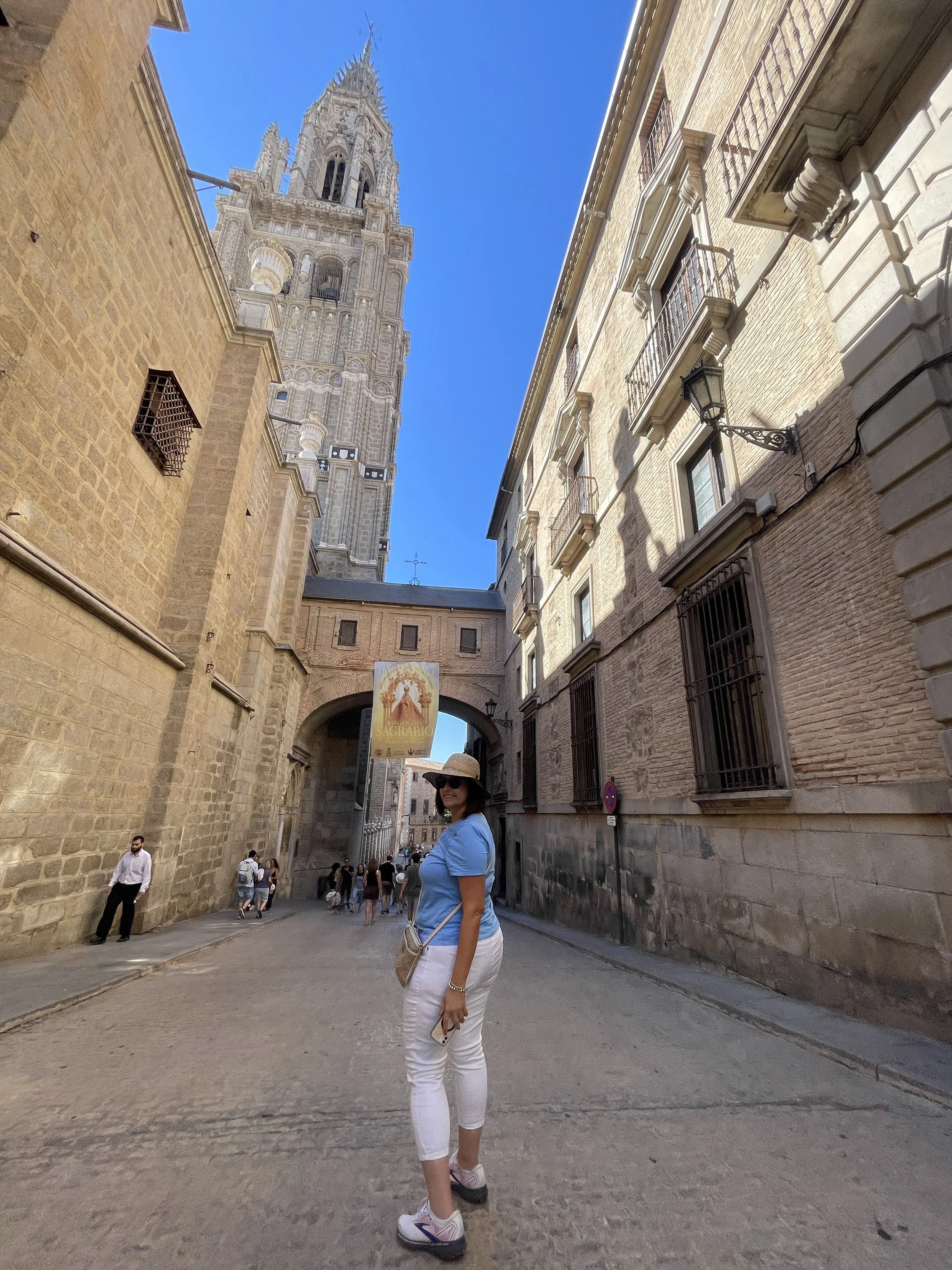 A woman wearing a blue shirt, white pants, and a sun hat standing on a cobblestone street in front of historic buildings and a tall clock tower in a European city.