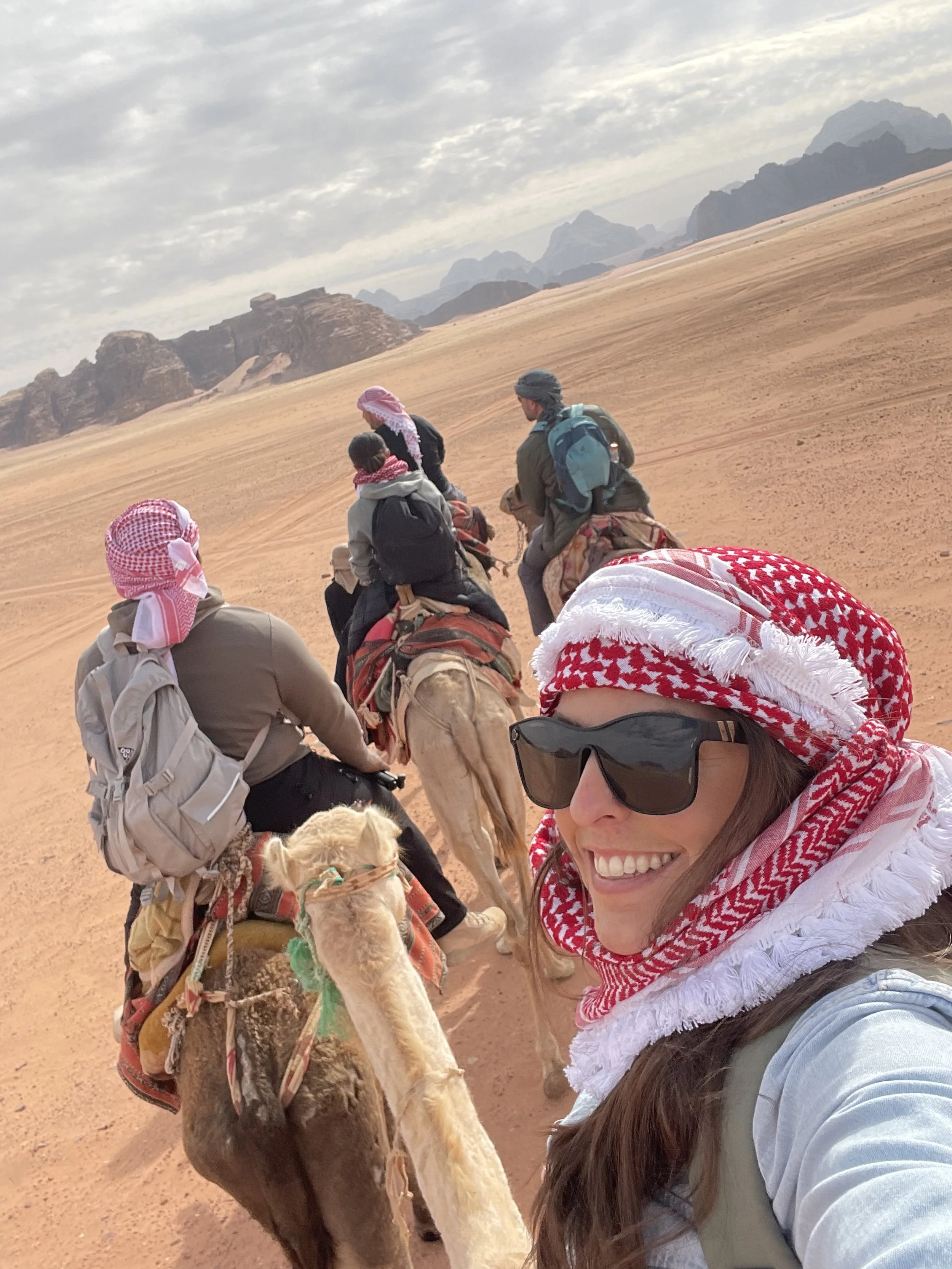A woman taking a selfie with a group of people and camels in a desert landscape.