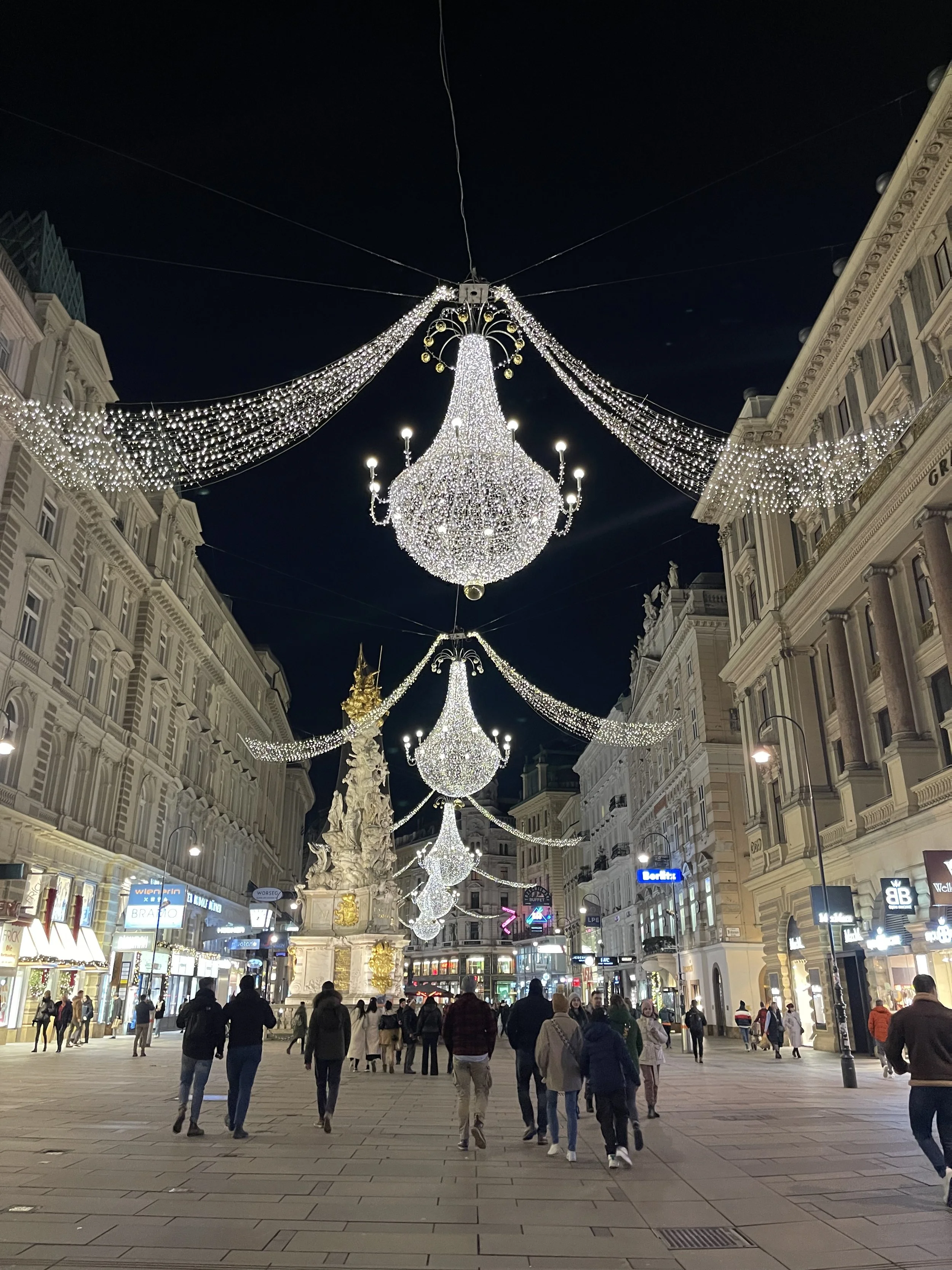 Street decorated with large chandeliers and hanging string lights during nighttime with people walking.
